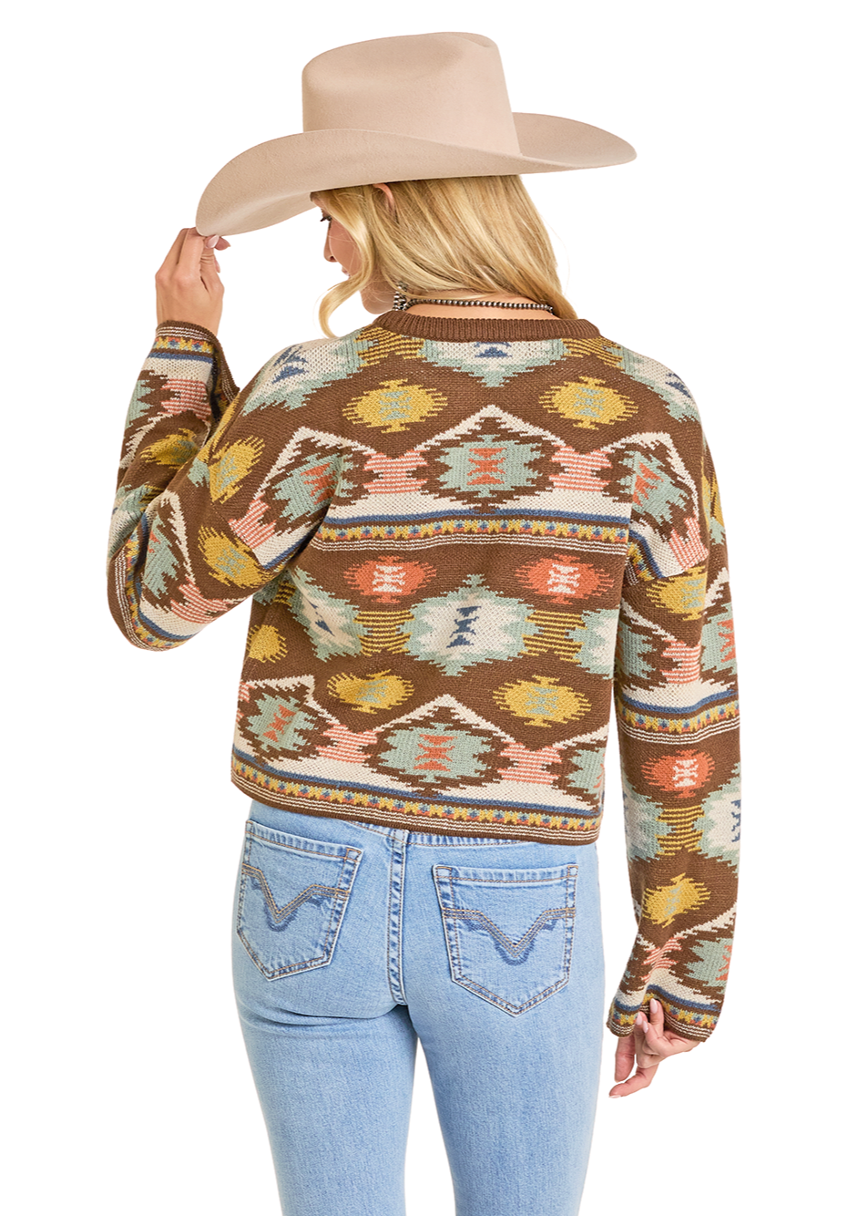 A woman in a beige cowboy hat and light blue jeans faces away, wearing the Rock & Roll Denim Aztec Cropped Sweater with a colorful Southwestern pattern in brown, yellow, blue, and pink. She holds her hat's brim with one hand.
