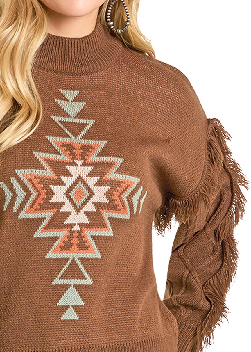A woman models the Rock & Roll Denim Aztec Knit Fringe Sweater, featuring a geometric tribal pattern in white, orange, and teal with fringe along the sleeves. Only her lower face and upper body are visible.