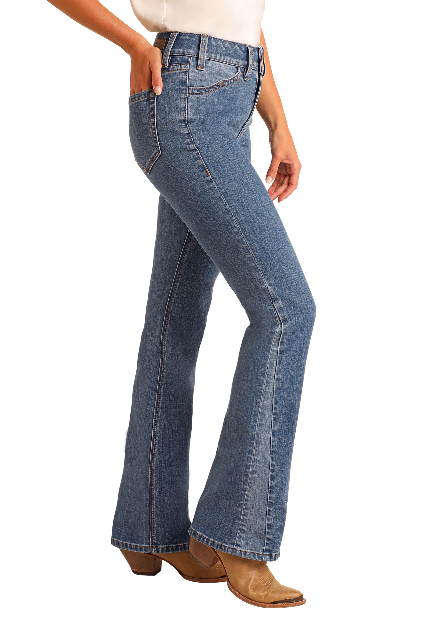 A woman in Rock & Roll Denim High Rise Two-Tone Detail Bootcut Jeans, a white top, and tan heeled boots stands sideways with one hand in her pocket against a plain light background.