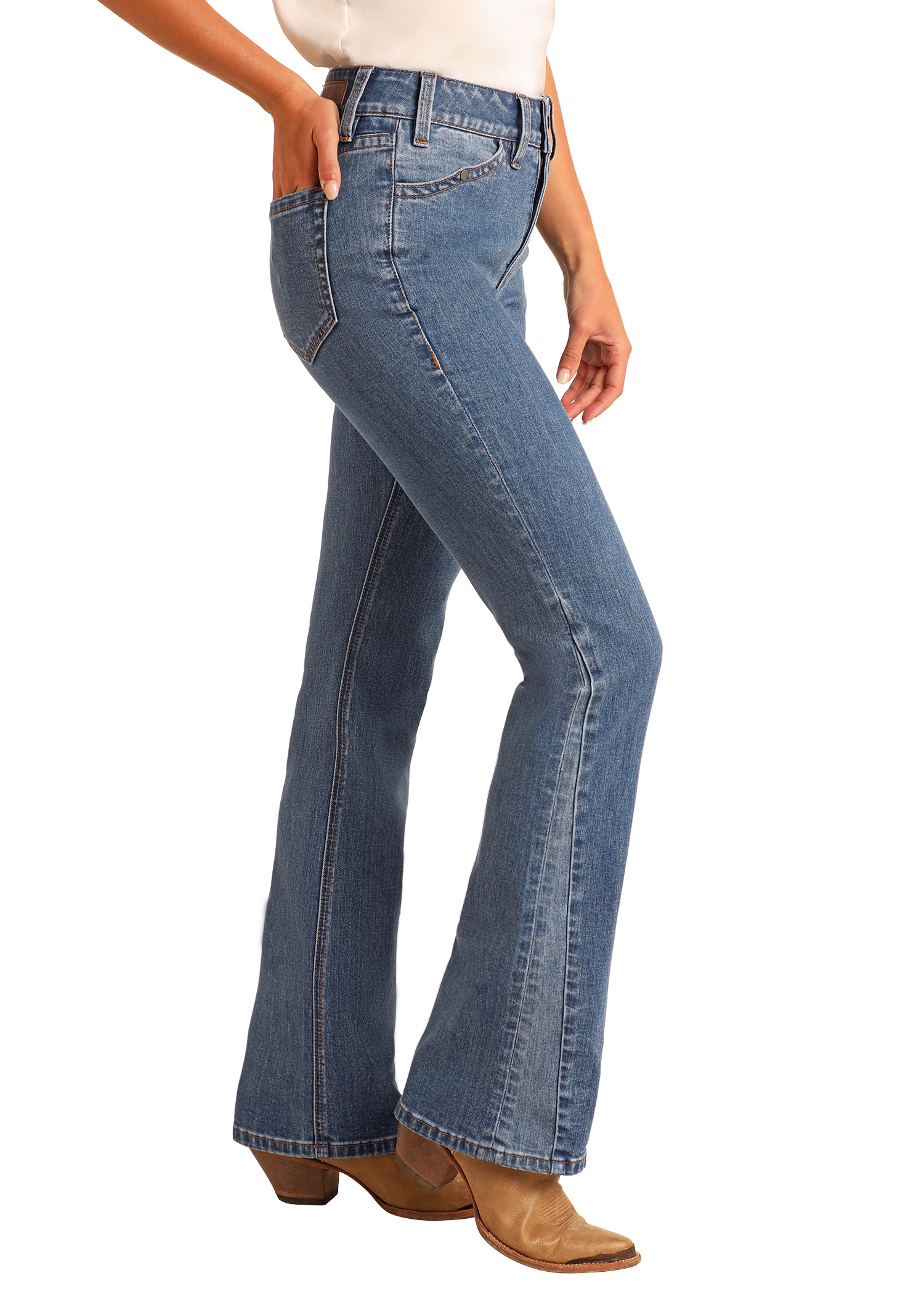 A woman in Rock & Roll Denim High Rise Two-Tone Detail Bootcut Jeans, a white top, and tan heeled boots stands sideways with one hand in her pocket against a plain light background.