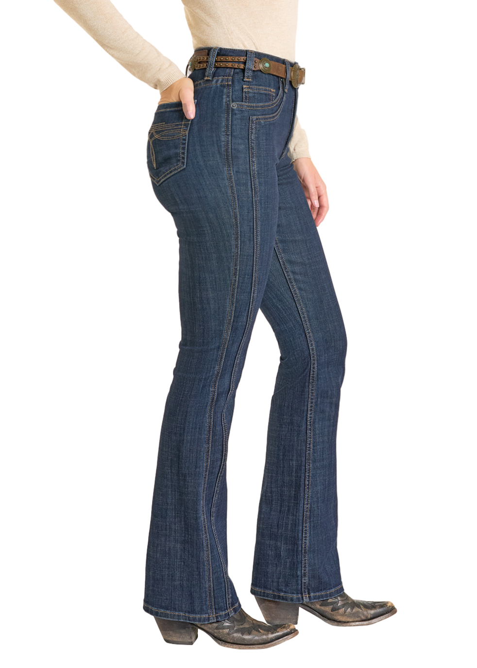 A person wearing Rock & Roll Denim High Rise Saddle Stitch Bootcut Jeans, a tan long-sleeve shirt, brown belt, and pointed-toe cowboy boots stands sideways with one hand in their pocket against a white background.