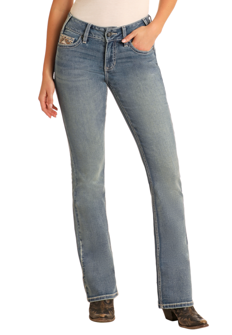 A woman wears Rock & Roll Denim Mid Rise Steer Head Embroidered Bootcut Jeans, a beige top, and cowboy boots, posing with one hand in her pocket against a plain background.