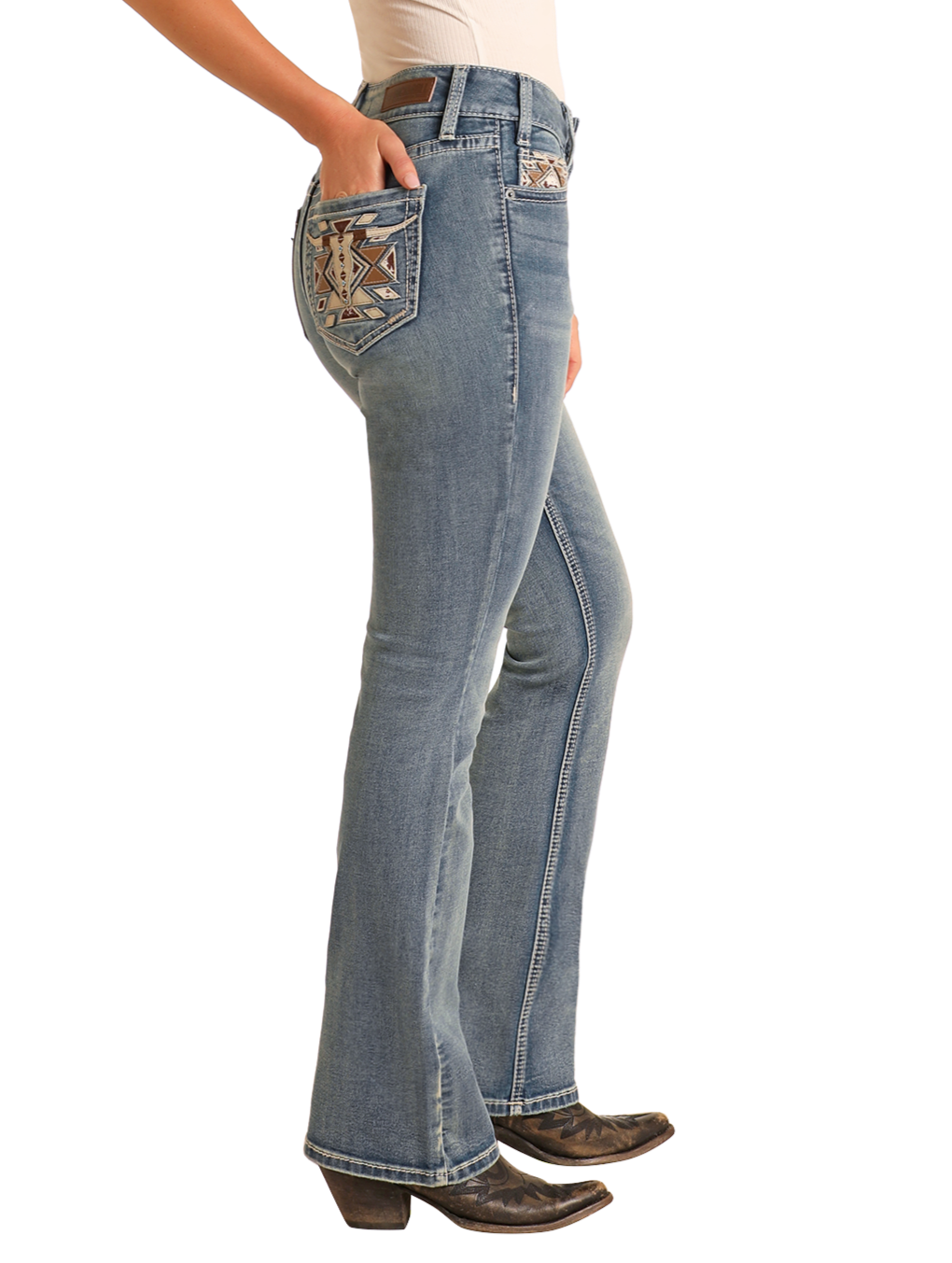 A person wears Rock & Roll Denim Mid Rise Steer Head Embroidered Bootcut Jeans with a white tucked-in shirt and black heeled boots, hands in pockets, standing against a light background.