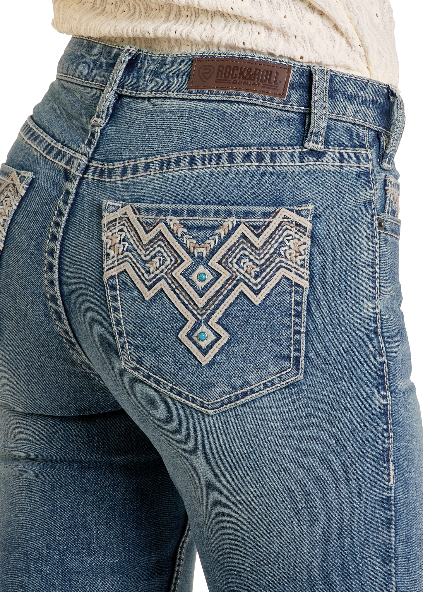 Close-up of a person wearing Rock & Roll Denim Mid Rise Aztec Embroidered Bootcut Jeans, featuring intricate white and turquoise back pocket designs and a branded leather patch on the waistband.