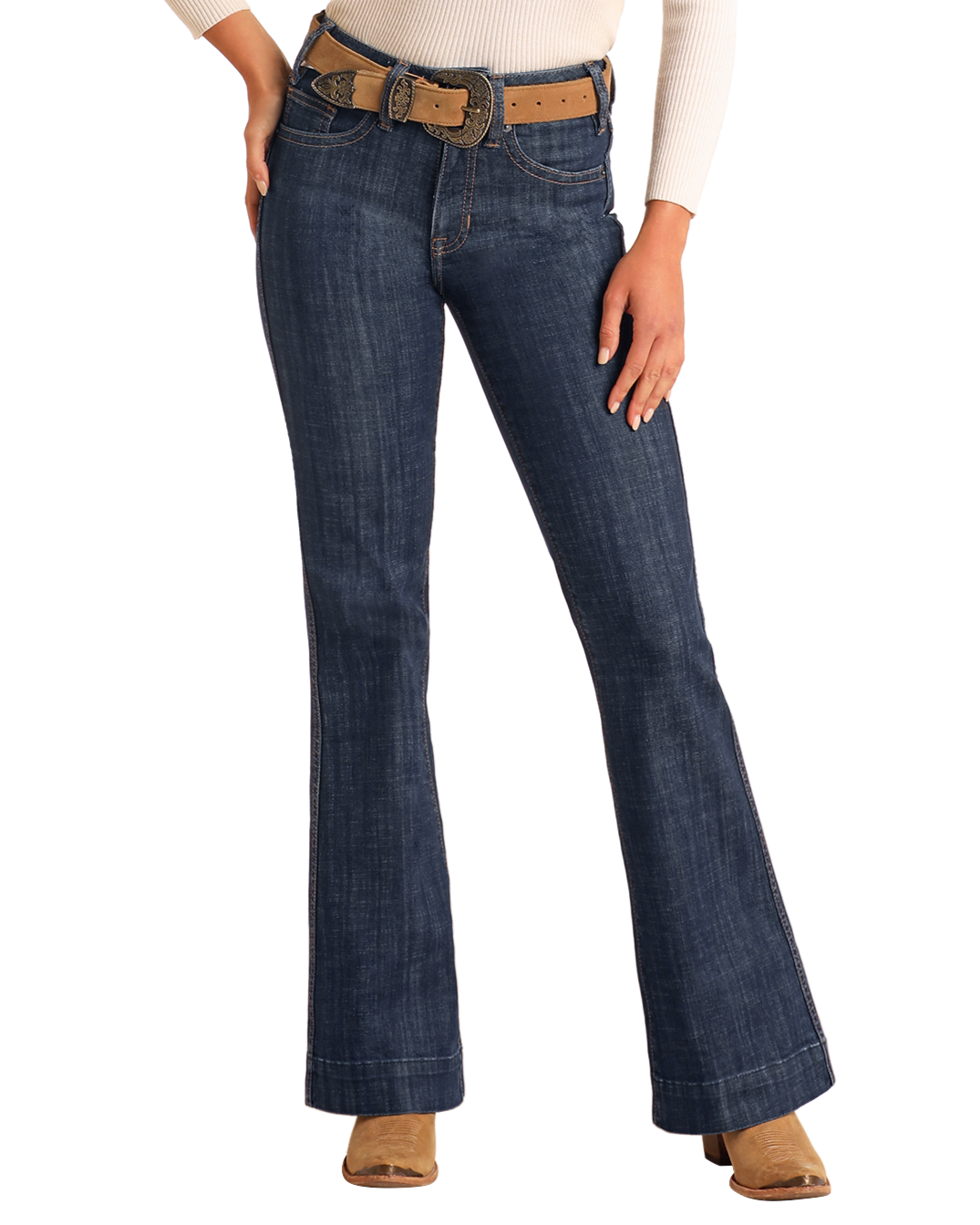 A person stands against a plain background, wearing Rock & Roll Denim’s High Rise Welt Pocket Trouser Jeans in dark blue, styled with a tan belt, cream long-sleeve top, and tan suede boots, one hand on their hip.