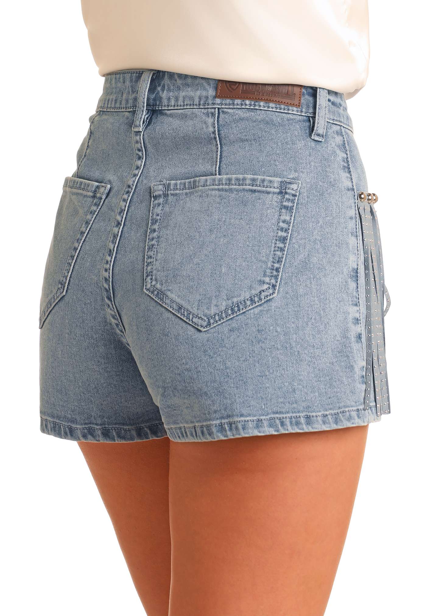 A person shown waist down from the back wears Rock & Roll Denim Extra Stretch Studded Fringe Shorts in light blue, featuring two back pockets and a small decorative chain, adding Western charm with subtle detailing.