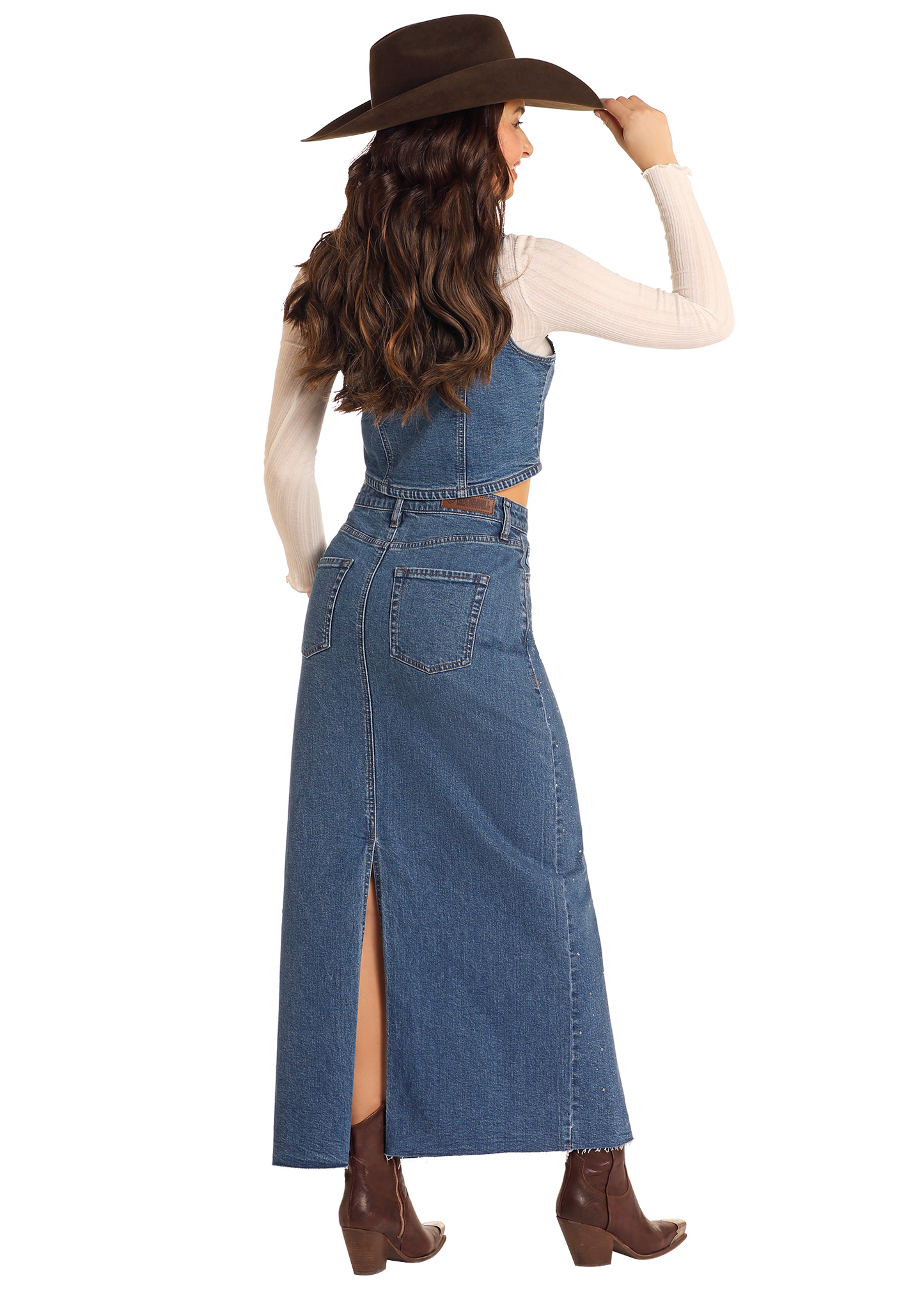 A woman stands looking to the side, wearing a brown hat, beige long-sleeve top, crop denim top, brown ankle boots, and the Rock & Roll Denim Studded Denim Maxi Skirt featuring a back slit.