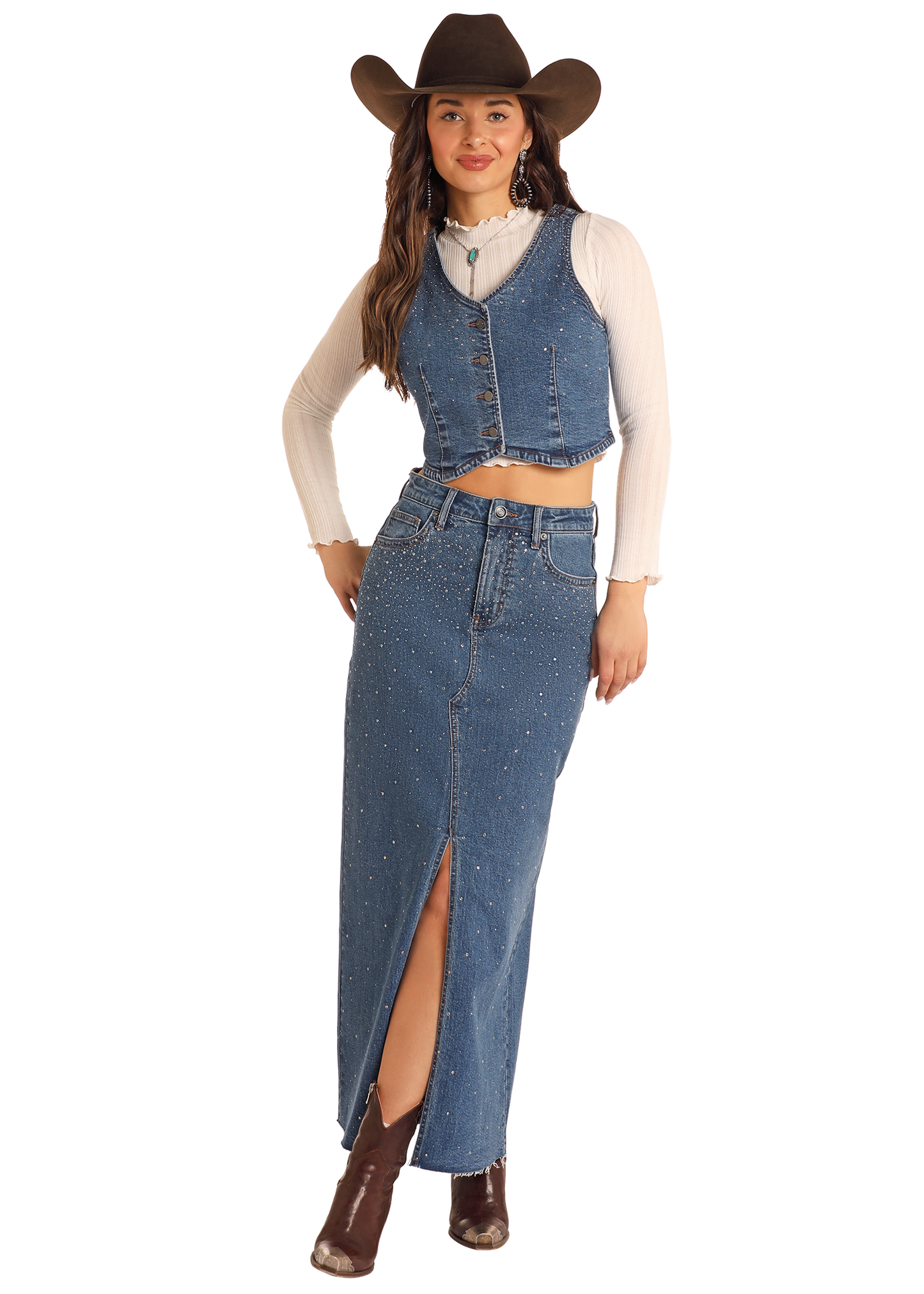 A woman stands confidently against a plain background in a brown cowboy hat, cream long-sleeve top, denim vest, and the Rock & Roll Denim Studded Denim Maxi Skirt with a front slit, accessorized with a necklace and brown ankle boots.
