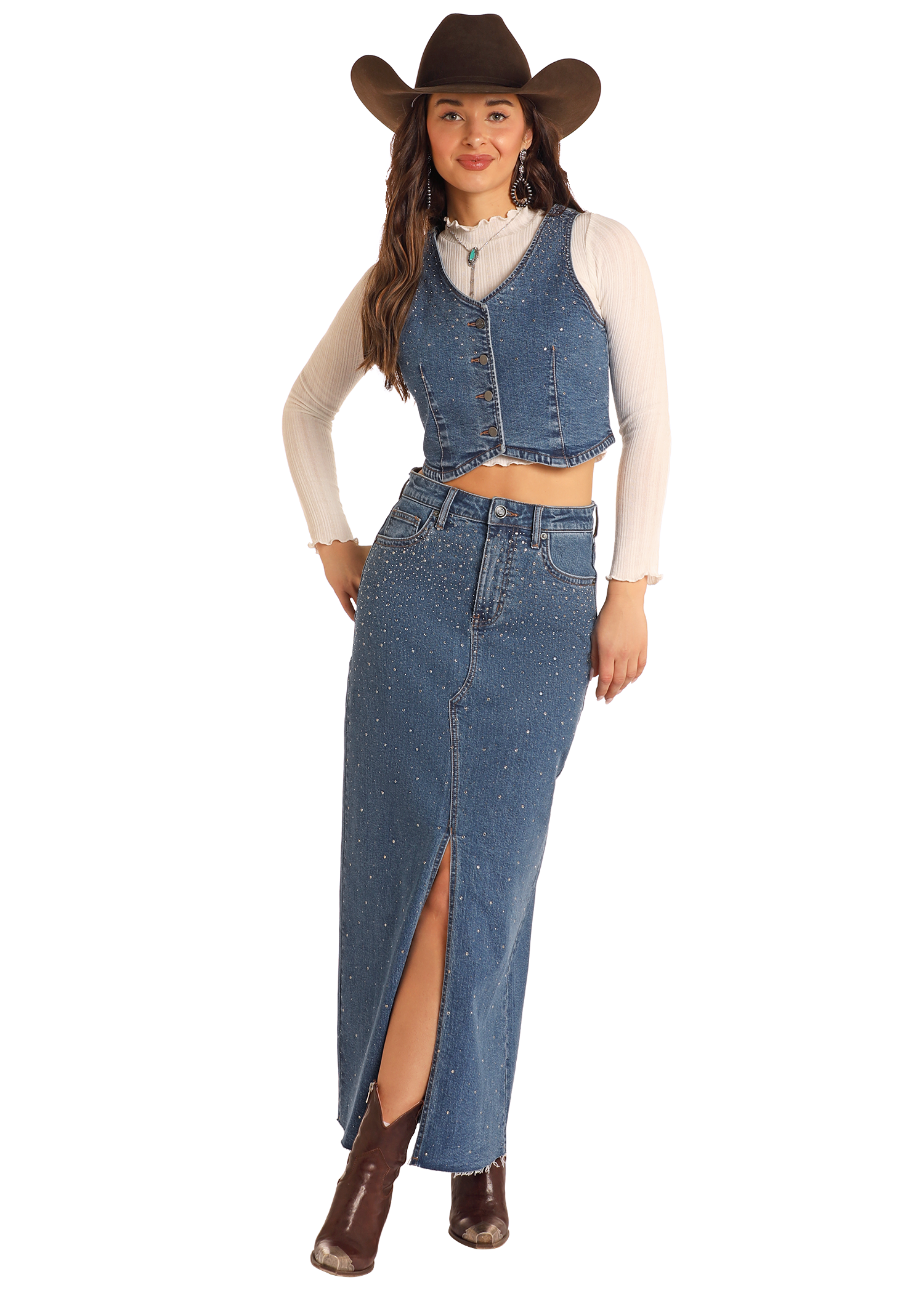 A woman stands confidently against a plain background in a brown cowboy hat, cream long-sleeve top, denim vest, and the Rock & Roll Denim Studded Denim Maxi Skirt with a front slit, accessorized with a necklace and brown ankle boots.