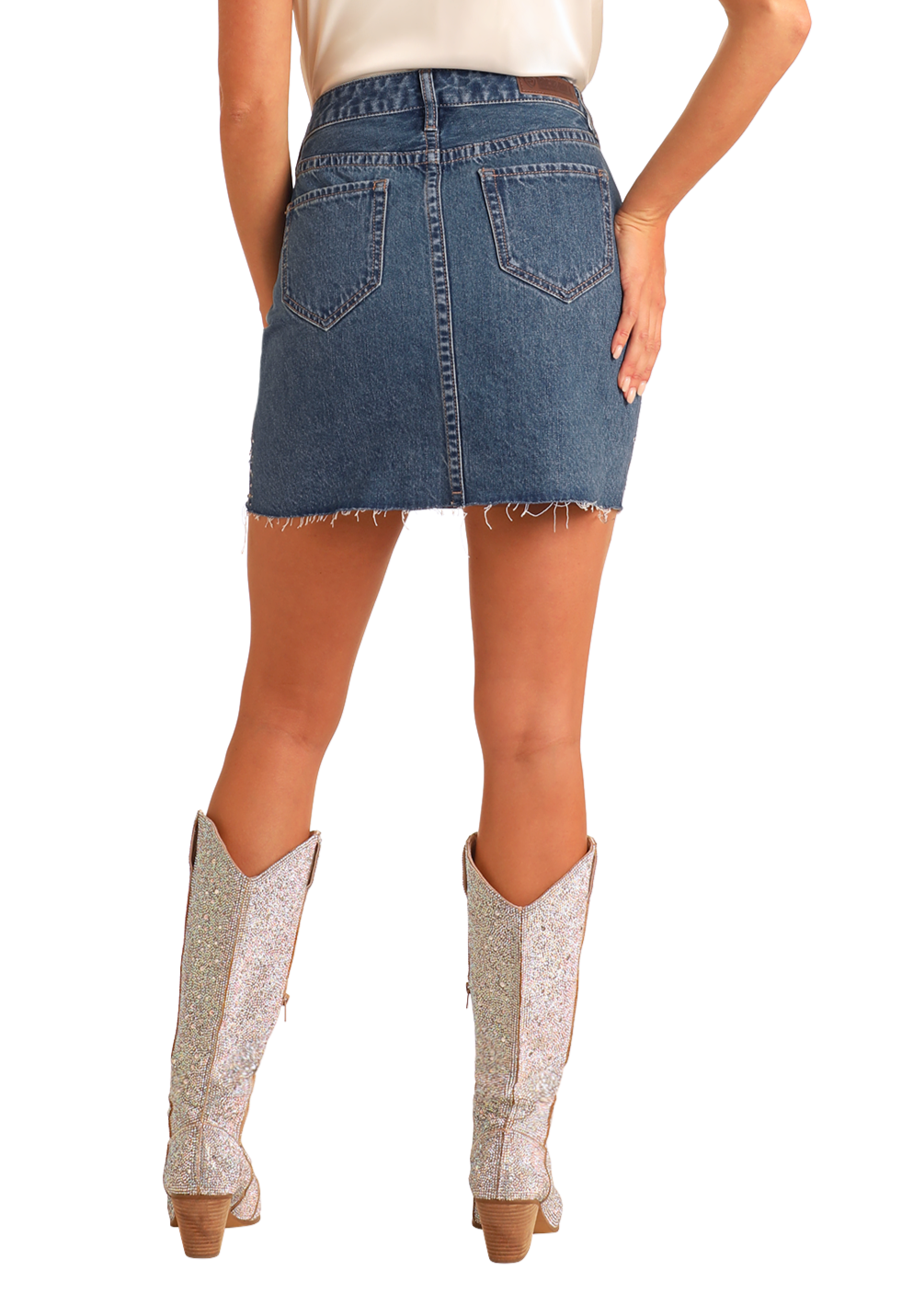 A woman stands facing away, hand on hip, wearing a Rock & Roll Denim Distressed Studded Denim Mini Skirt with frayed hem, a light top, and sparkly knee-high cowboy boots with wooden heels.