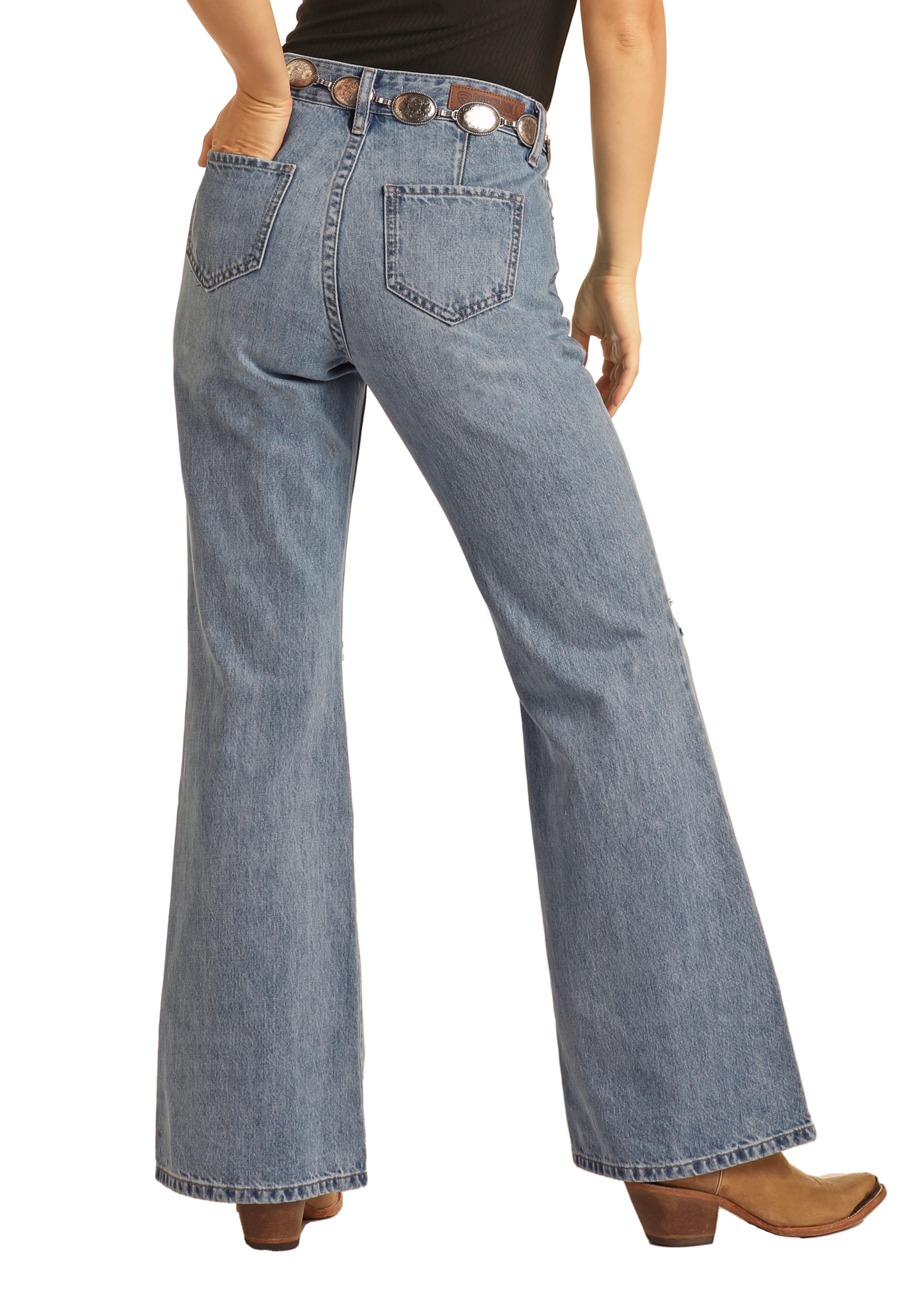 A person stands with their back to the camera, wearing Rock & Roll Denim High Rise Distressed Relaxed Wide Flare Jeans in light blue, paired with a black top, dark belt with large round buckles, and brown heeled shoes.