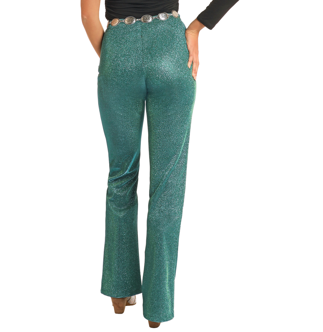 A person faces away, one hand on their hip, wearing Rock & Roll Denim’s turquoise Iridescent Pants with a decorative belt, styled with a long-sleeved black top and heeled shoes.