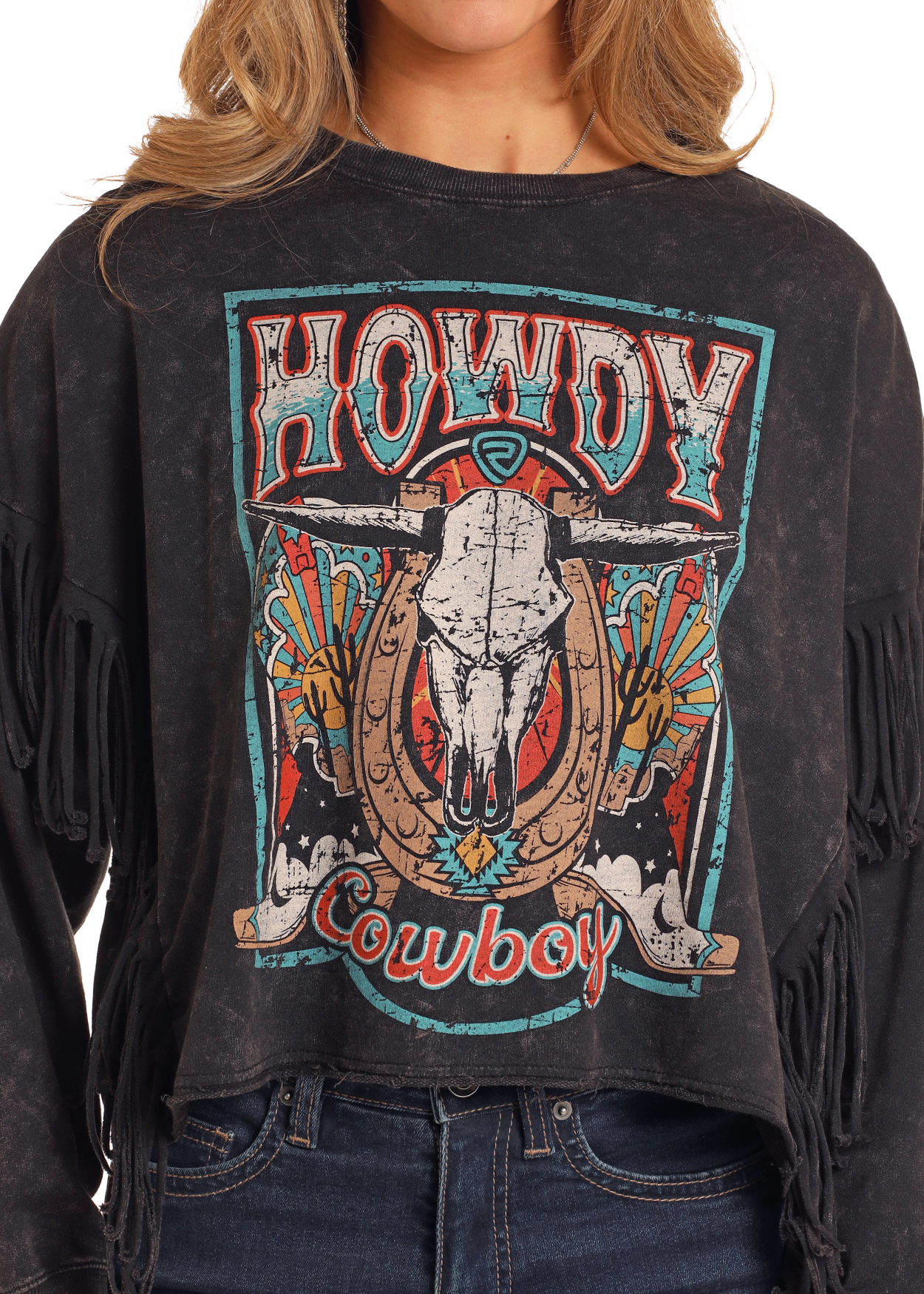 A woman, showing only her lower face and torso, wears the Rock & Roll Denim Howdy Cowboy Fringe Pullover featuring a colorful “Howdy Cowboy” graphic with a steer skull, horseshoes, and southwestern motifs.