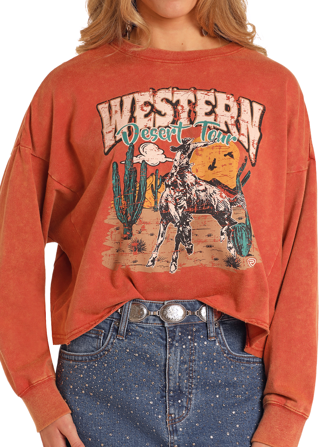 A woman wears the Rock & Roll Denim Western Desert Tour Pullover, featuring a horse, cacti, and mountains graphic. She styles the faded red sweatshirt with high-waisted blue jeans and a silver belt for an effortlessly cool vibe.