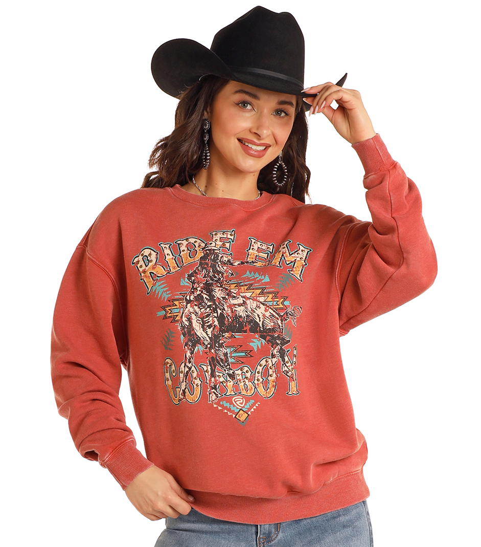 A woman in a black cowboy hat and bold earrings poses, smiling, in the Rock & Roll Denim Studded Ride 'Em Cowboy Oversized Pullover featuring a sparkling graphic of a cowboy on horseback and “Ride ’Em Cowboy” text.