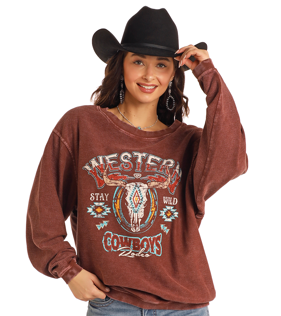A smiling woman in a black cowboy hat and hoop earrings models the Rock & Roll Denim Western Cowboys Ribbed Pullover, featuring a loose fit and Western Cowboys with bull graphic on the front.