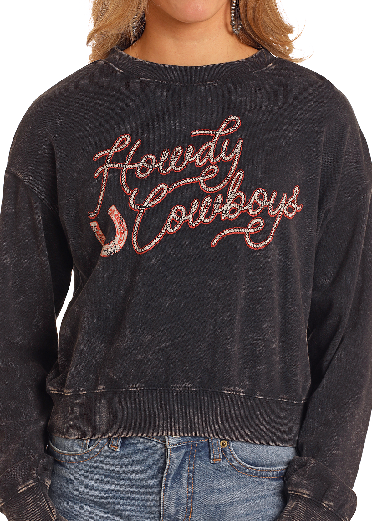 A person wears the Rock & Roll Denim Howdy Cowboys Cropped Pullover, a black acid-wash sweatshirt with red and white rope-style lettering, paired with blue jeans for a bold Western graphic look.