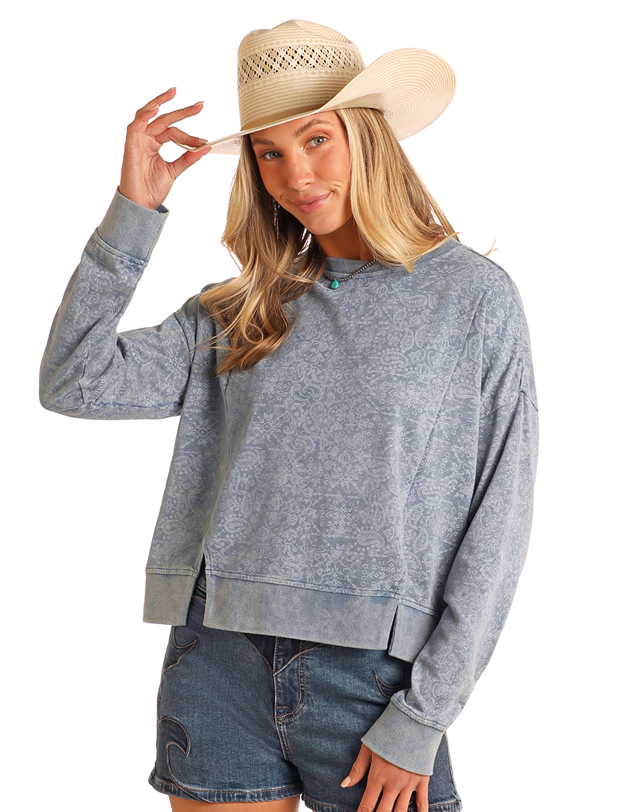 A woman wearing a relaxed-fit Rock & Roll Denim Bandana Print Pullover, denim shorts, and a beige cowboy hat smiles while holding her hat's brim against a plain white background.
