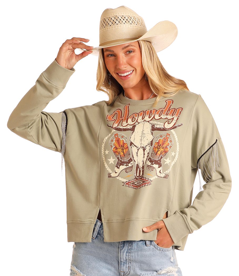 A smiling woman in a straw cowboy hat and ripped denim shorts poses against a plain background, wearing the Rock & Roll Denim Chain Fringe Howdy Graphic Pullover and touching her hat.
