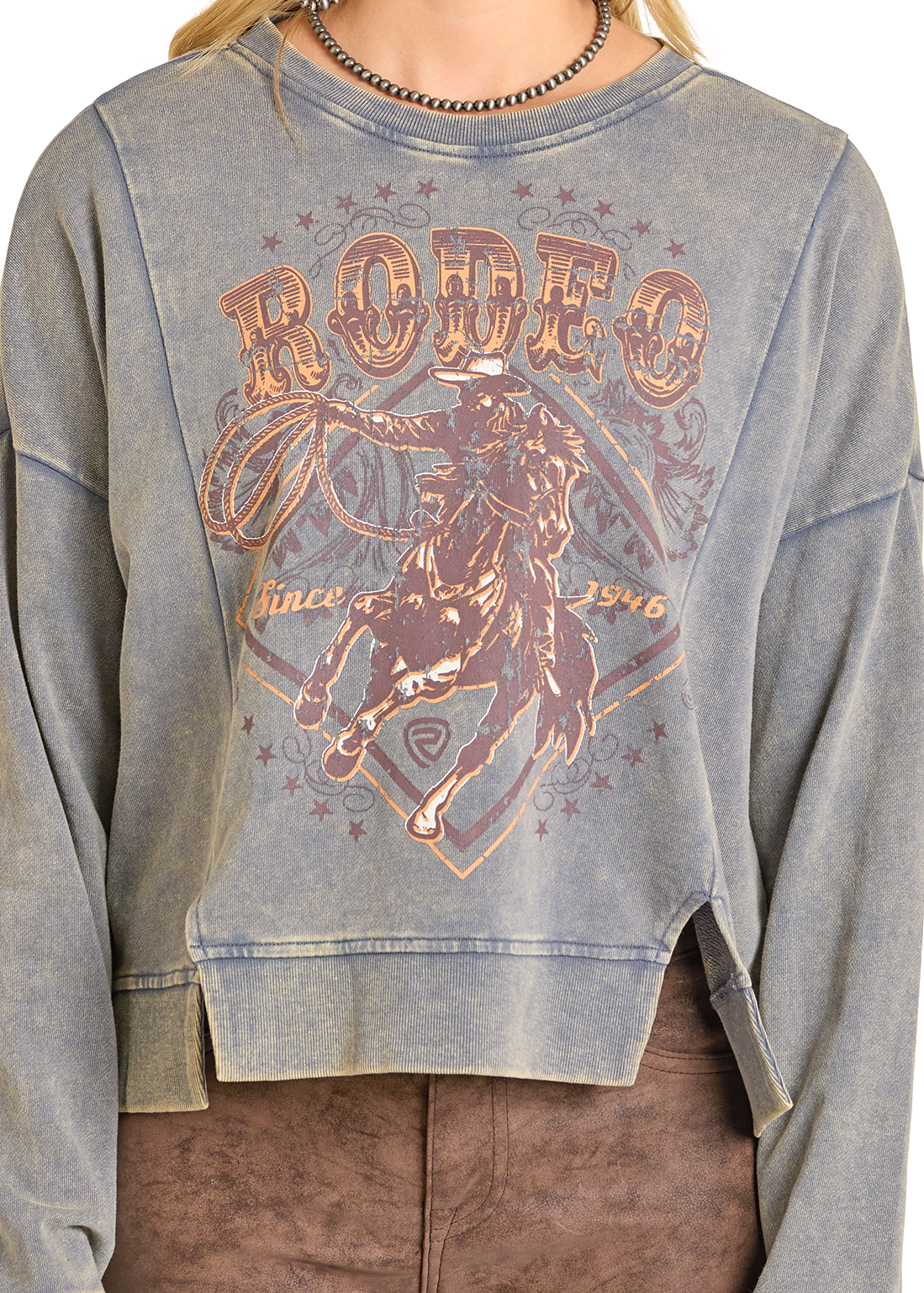 A person wears the Rock & Roll Denim Rodeo Graphic Print Pullover, a faded blue cropped top with a vintage cowboy and RODEO graphic, raw hems, and decorative text, styled with brown pants for a relaxed vibe.