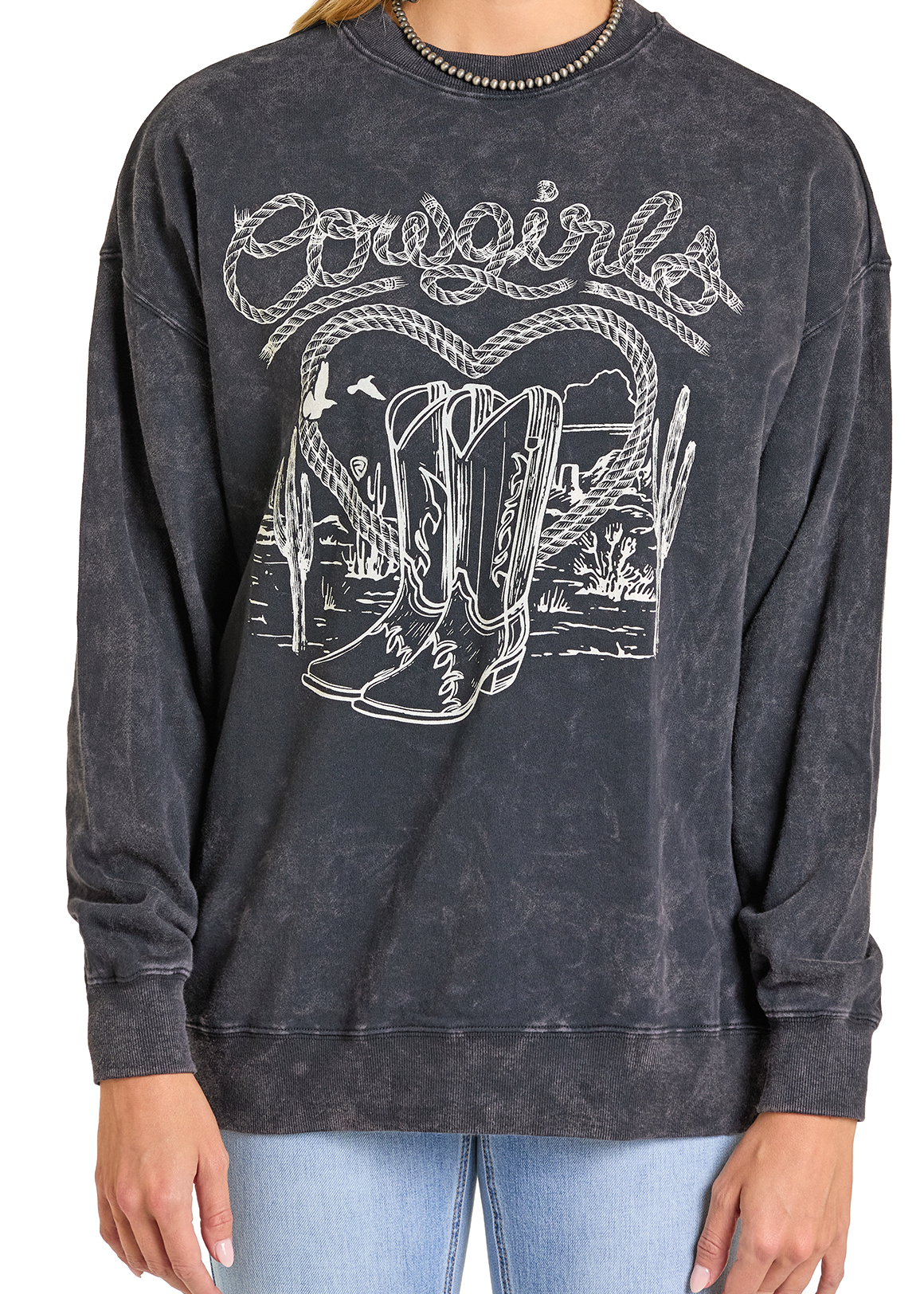 Someone wears the Rock & Roll Denim Cowgirl Graphic Pullover, a black mineral wash top with cowboy boots, cacti, mountains, and “Cowgirls” in bold script, styled with light blue jeans.