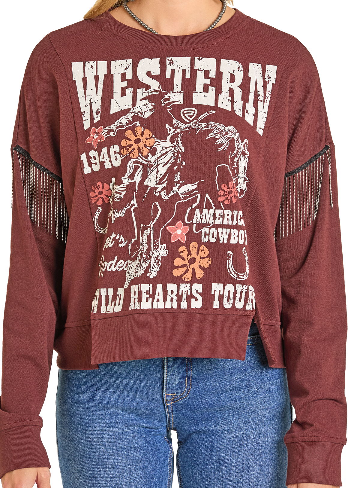 A person wears the Chain Fringe Western Graphic Pullover by Rock & Roll Denim, featuring fringe on the arms and a graphic with "WESTERN 1946 American Cowboy Rodeo Wild Hearts Tour," plus a horse, rider, flowers, and a horseshoe.
