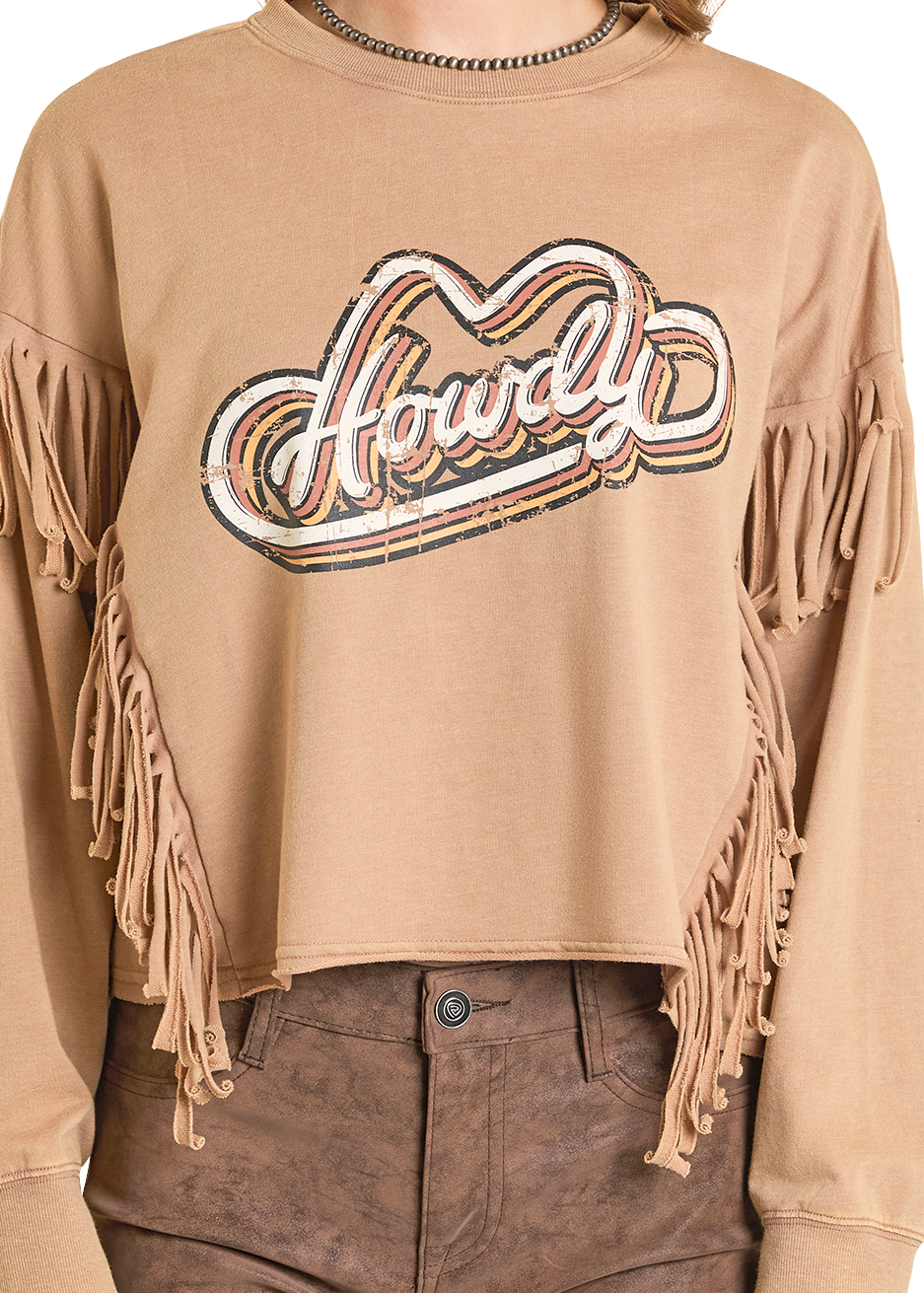 A person wears the tan Fringe Howdy Graphic Pullover by Rock & Roll Denim, featuring fringed sleeves and chest with a retro graphic, paired with high-waisted brown pants and a beaded necklace.