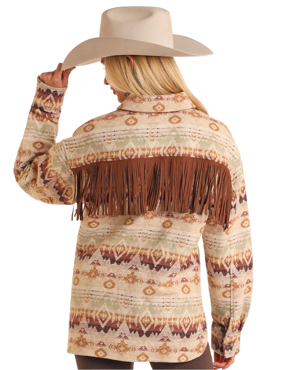 A woman in the Rock & Roll Denim Fringe Aztec Shacket, a 100% cotton Western-style shirt jacket with brown fringe, and a wide-brimmed beige cowboy hat faces away holding her hat—a timeless casual look.