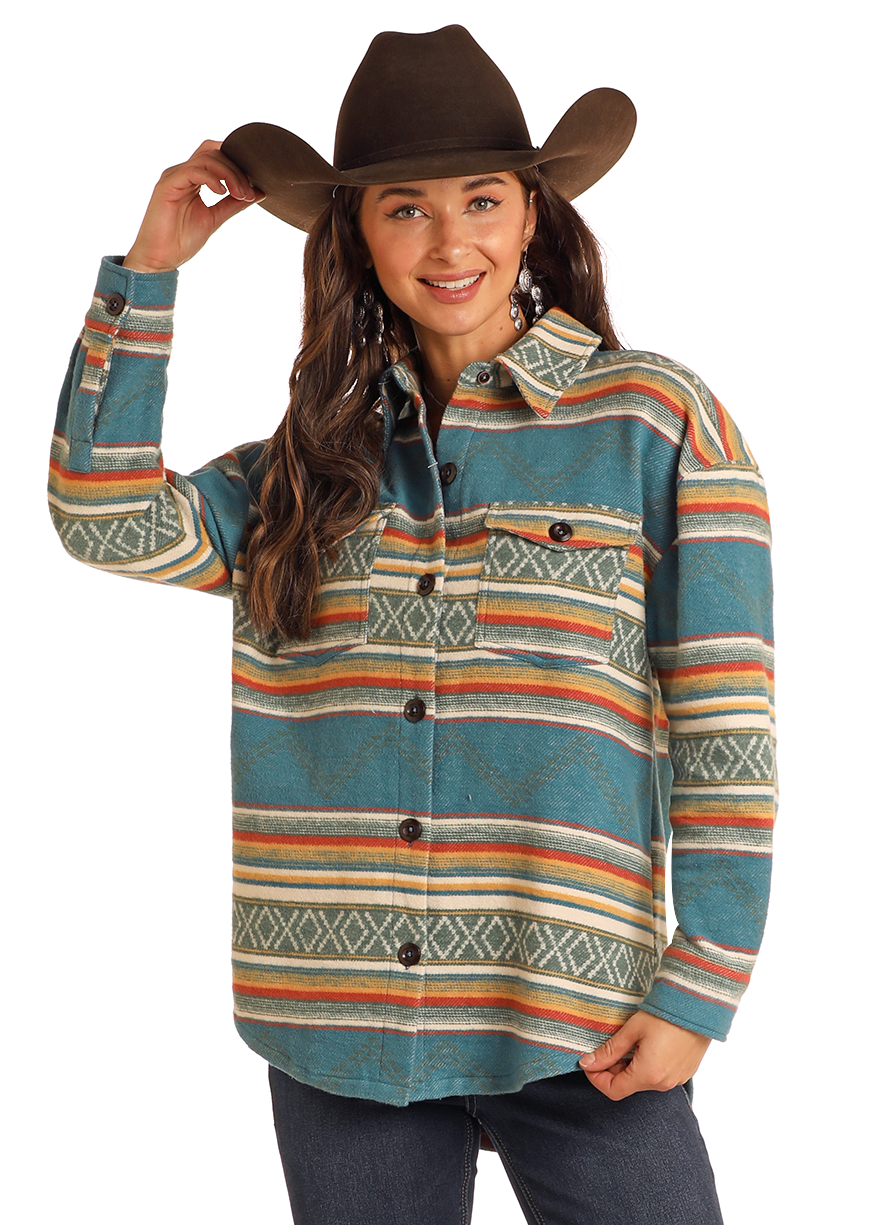 A smiling woman in a Rock & Roll Denim Turquoise Aztec Shacket, black cowboy hat, and dark jeans touches her hat brim while standing against a plain background.