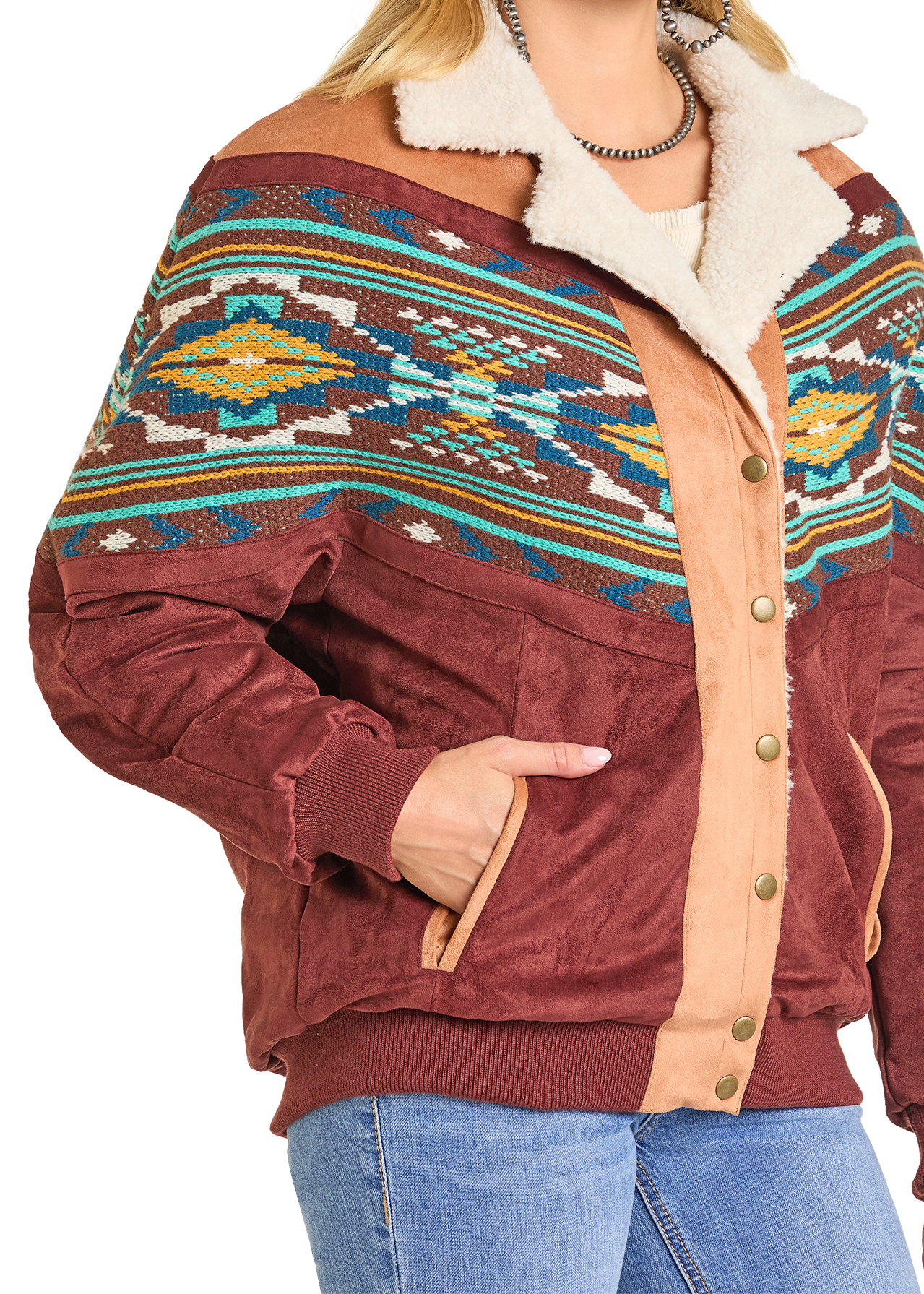 A person wears the Rock & Roll Denim Oversized Microsuede Bomber Jacket with a colorful Aztec pattern, faux fur collar, tan button placket, and blue jeans. One hand rests in the pocket of this bold brown bomber jacket.
