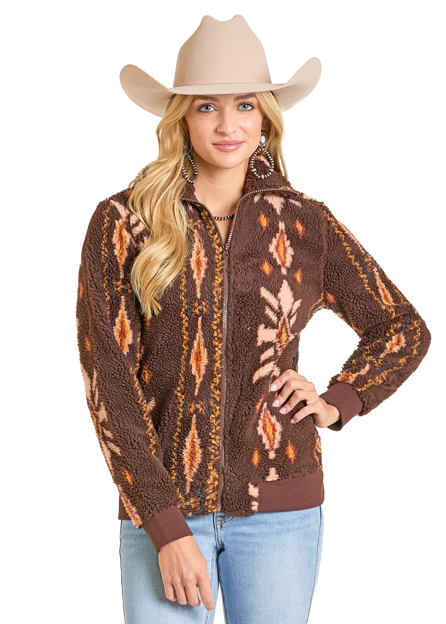 A woman wearing a beige cowboy hat, beaded earrings, light blue jeans, and the Rock & Roll Denim Brown Aztec Sherpa Jacket stands with one hand on her hip and smiles.