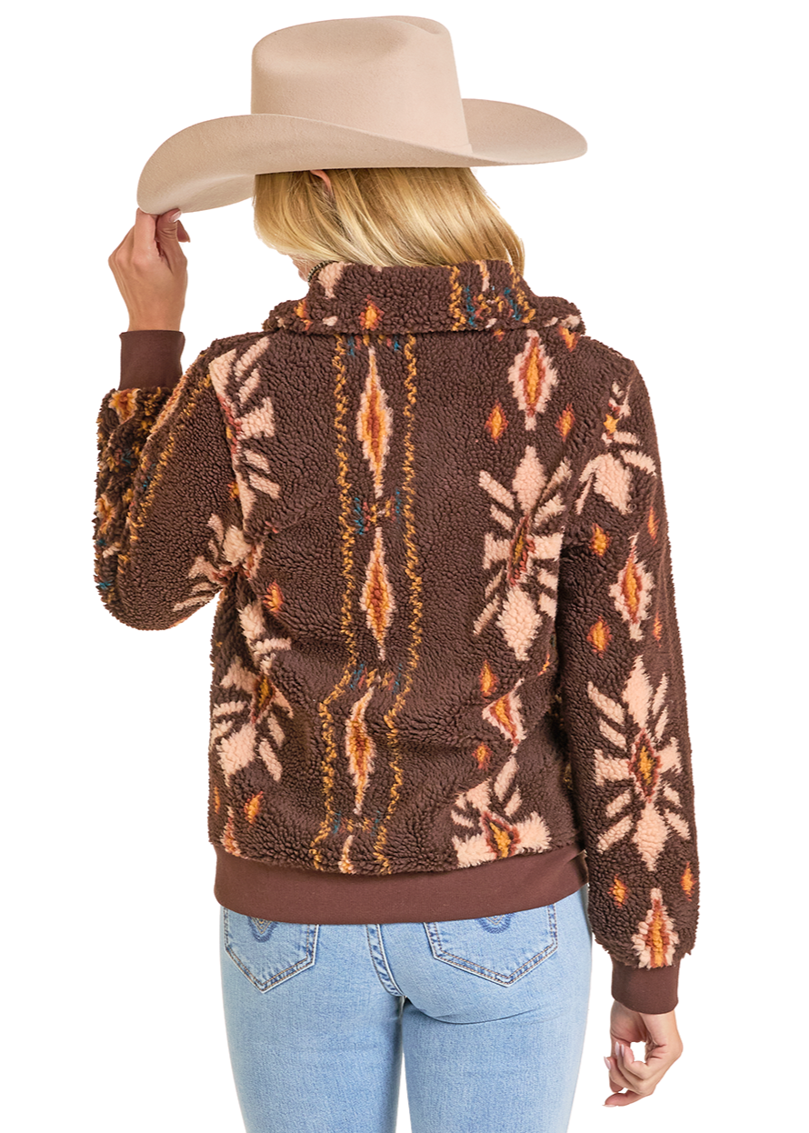 A woman in a beige cowboy hat and light blue jeans faces away, wearing the Rock & Roll Denim Brown Aztec Sherpa Jacket with an orange, tan, and cream pattern. She holds her hat brim with one hand.