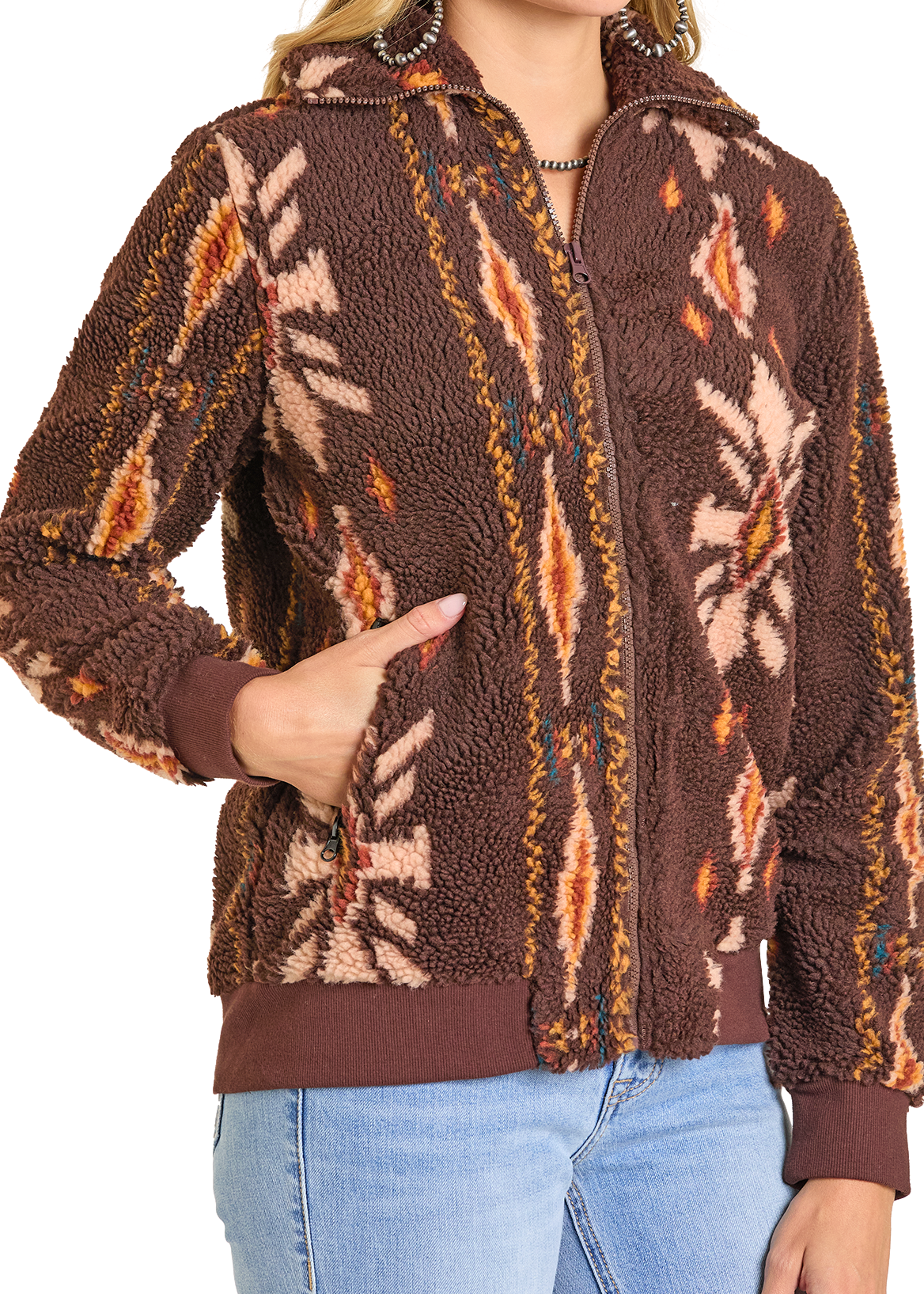 A person wears the Rock & Roll Denim Brown Aztec Sherpa Jacket, zipped up with one hand in a pocket. The women's jacket features ribbed cuffs and hem and pairs perfectly with light blue jeans.
