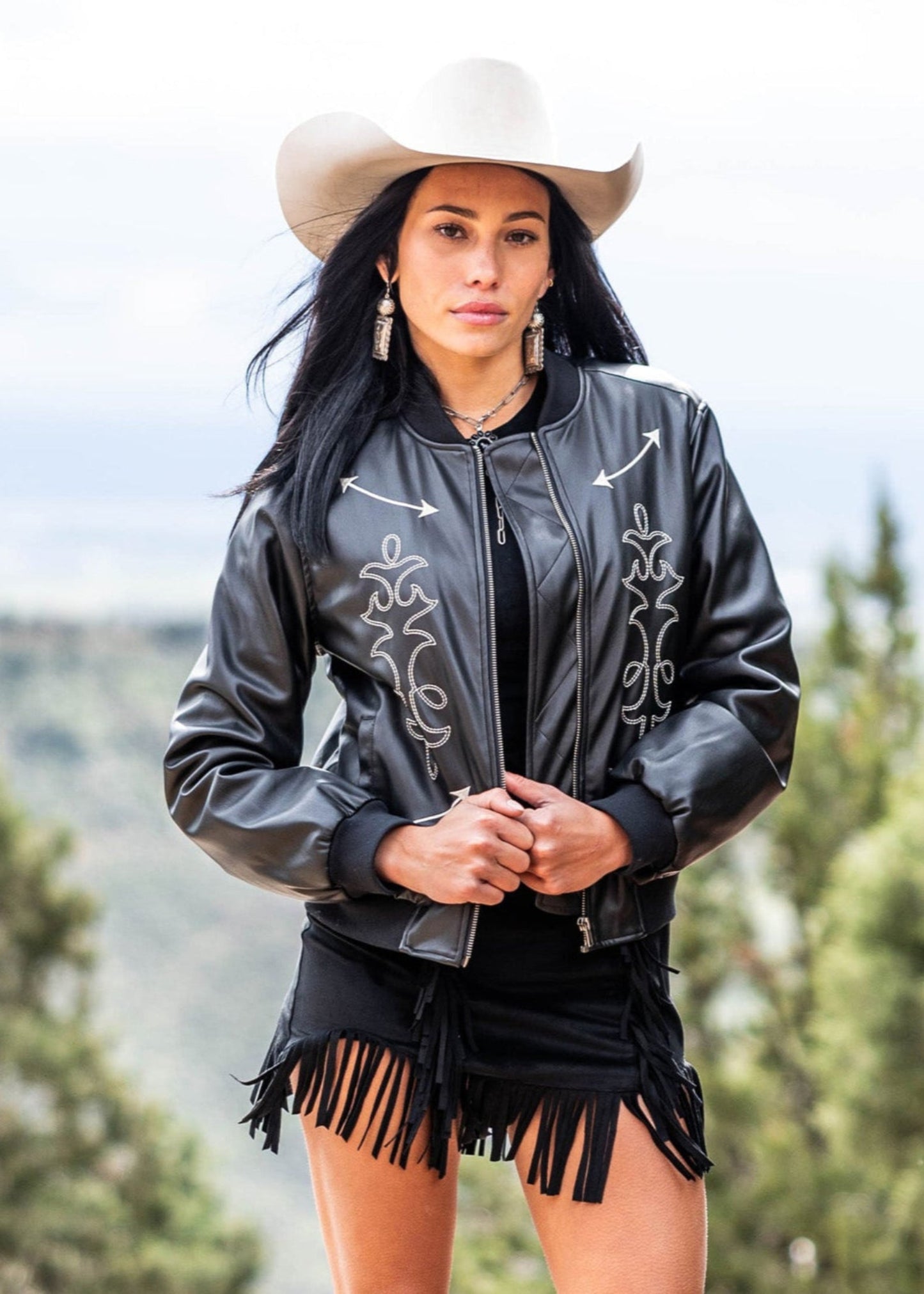 A woman wears a white cowboy hat, earrings, black fringe shorts, and the Rock & Roll Denim Pleather Bomber Jacket with chic appliqué and decorative stitching, standing outdoors against a blurred natural backdrop.