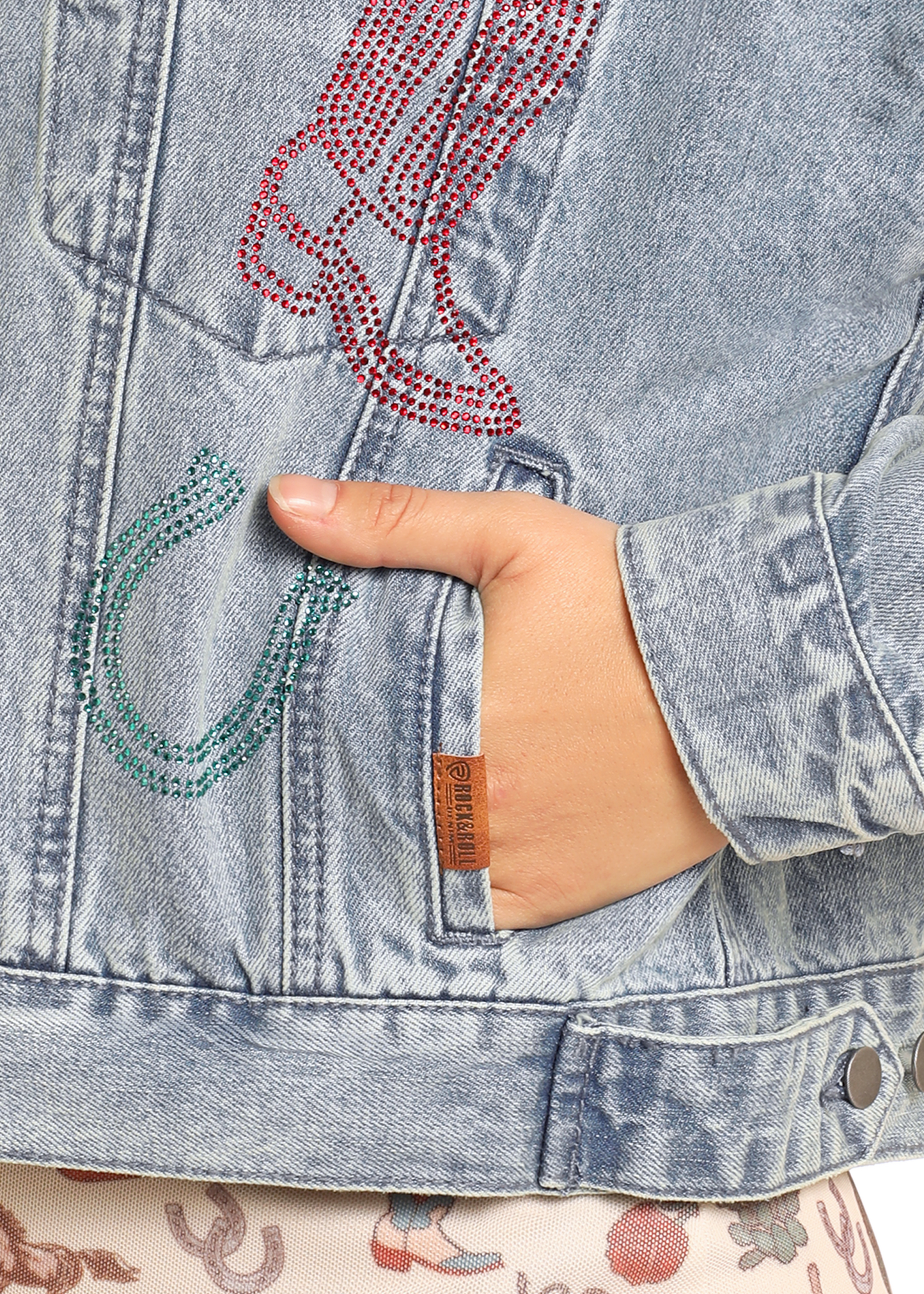 A hand rests in the pocket of the Rock & Roll Denim Studded Western Denim Jacket for women, featuring a light wash, rhinestone horseshoe and cowboy boot designs, stud accents, and a small brown tag near the pocket.