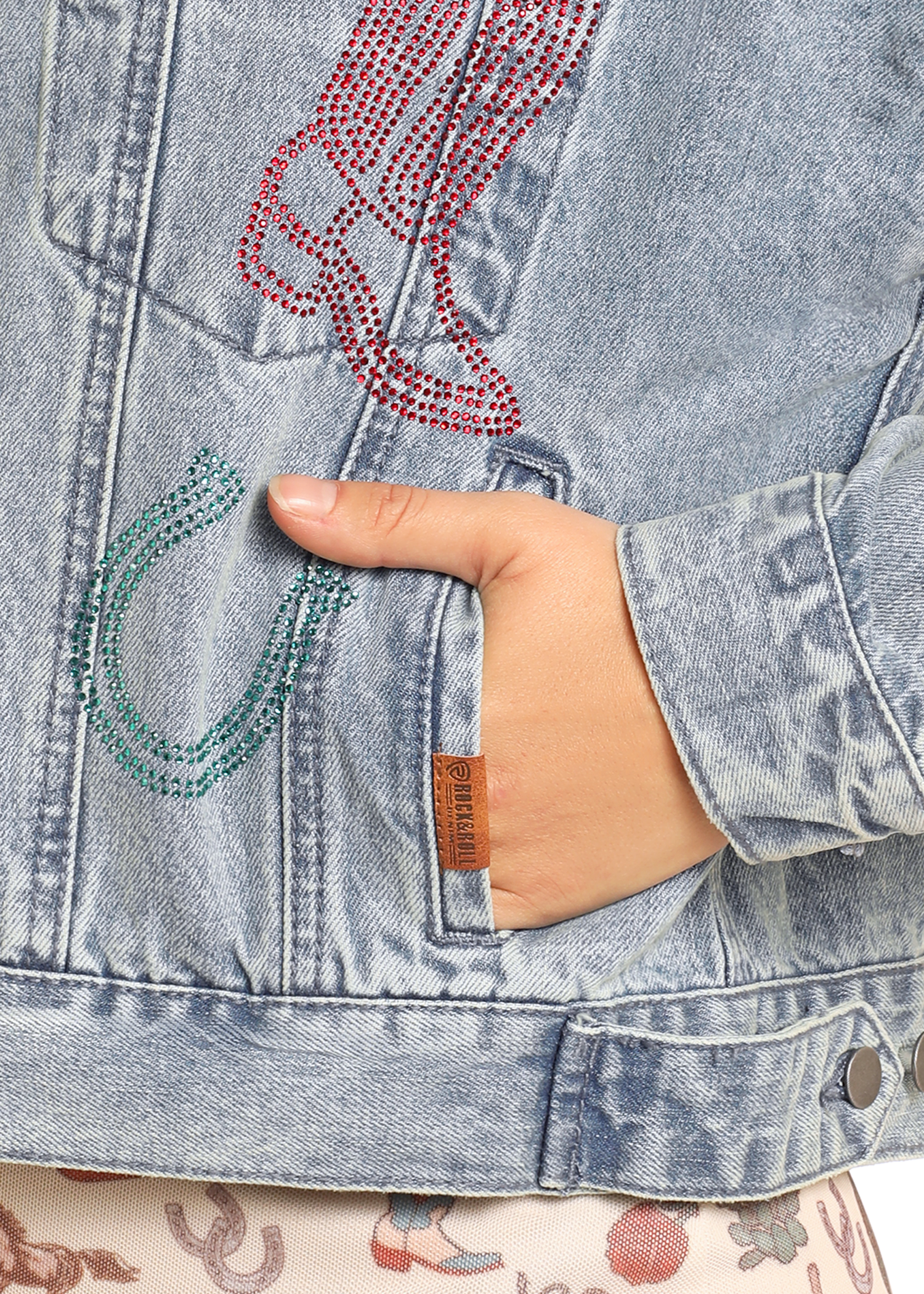 A hand rests in the pocket of the Rock & Roll Denim Studded Western Denim Jacket for women, featuring a light wash, rhinestone horseshoe and cowboy boot designs, stud accents, and a small brown tag near the pocket.