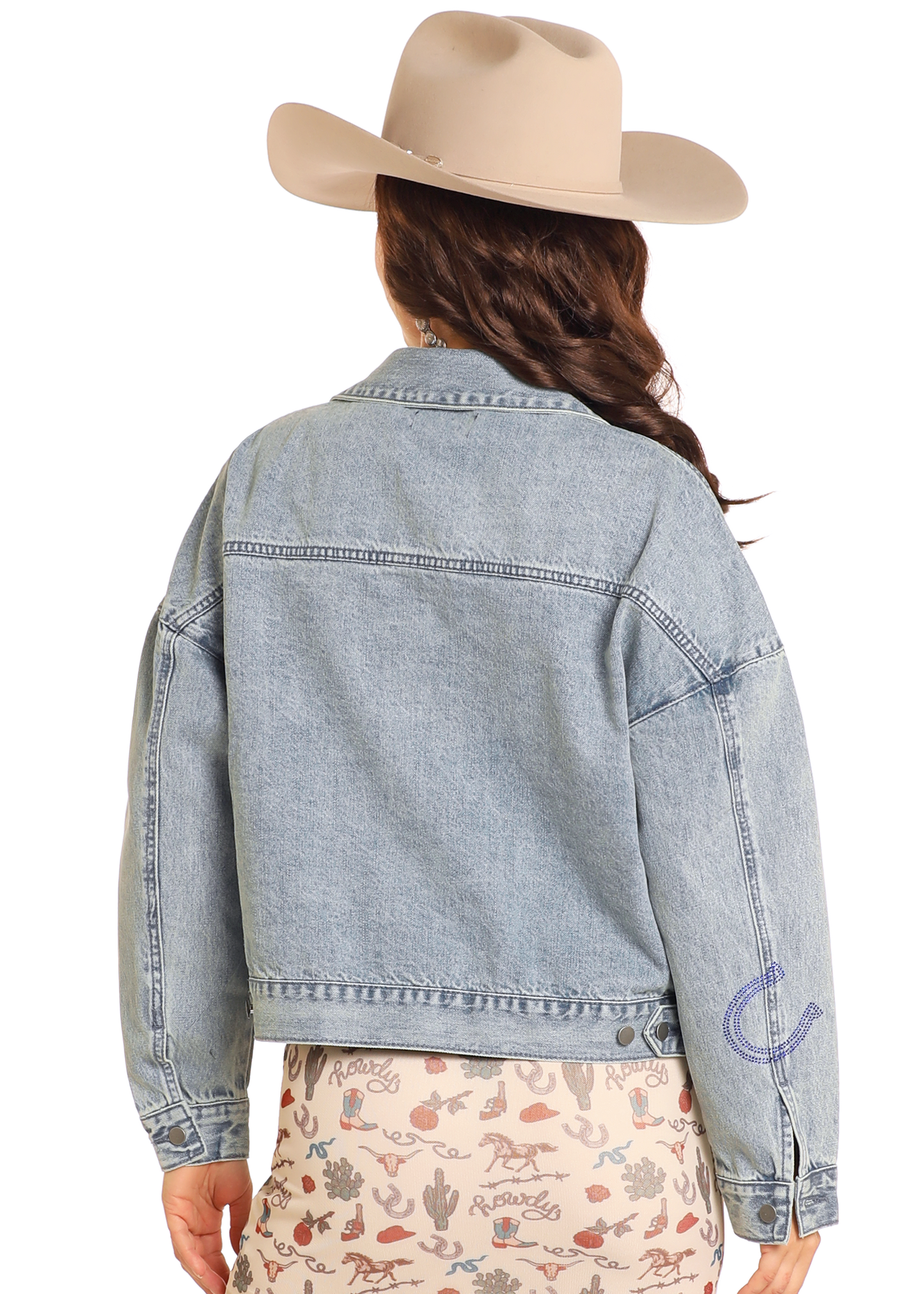 Shown from the back against a plain background, a person wears the Rock & Roll Denim Studded Western Denim Jacket, paired with a beige cowboy hat and a cream skirt featuring a western-themed print.