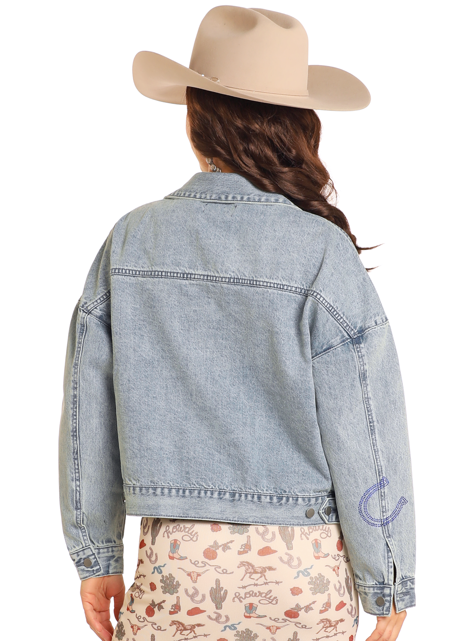 Shown from the back against a plain background, a person wears the Rock & Roll Denim Studded Western Denim Jacket, paired with a beige cowboy hat and a cream skirt featuring a western-themed print.
