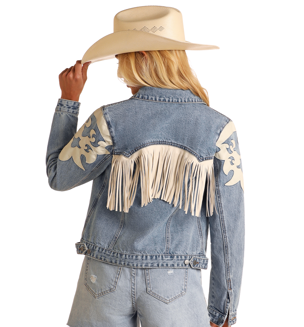 A woman wearing the Rock & Roll Denim Pleather Fringe Denim Jacket, featuring cream fringe and decorative patches, is paired with light denim shorts and a wide-brimmed cowboy hat, facing away from the camera.