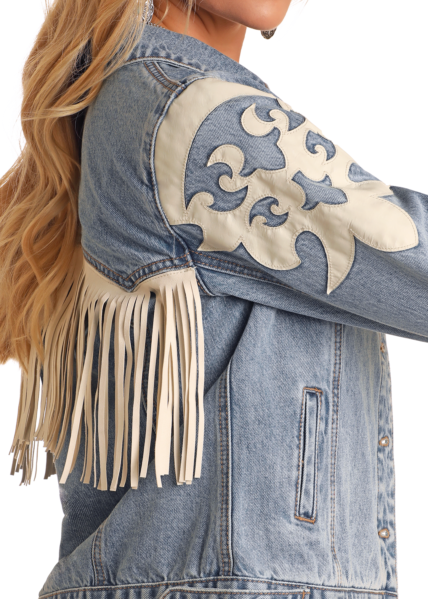 A woman with long blonde hair wears the Rock & Roll Denim Pleather Fringe Denim Jacket—a blue denim piece with tan pleather fringe and white appliqué designs on the shoulder and sleeve.