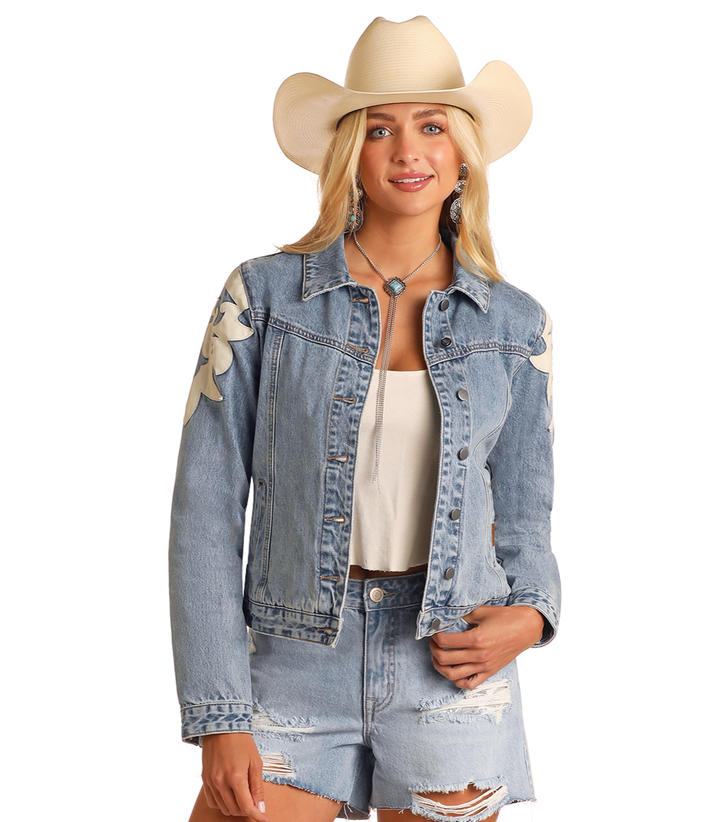 A woman smiles with one hand in her pocket, wearing the Rock & Roll Denim Pleather Fringe Denim Jacket over a white top and ripped denim shorts, paired with a white cowboy hat, against a plain background.