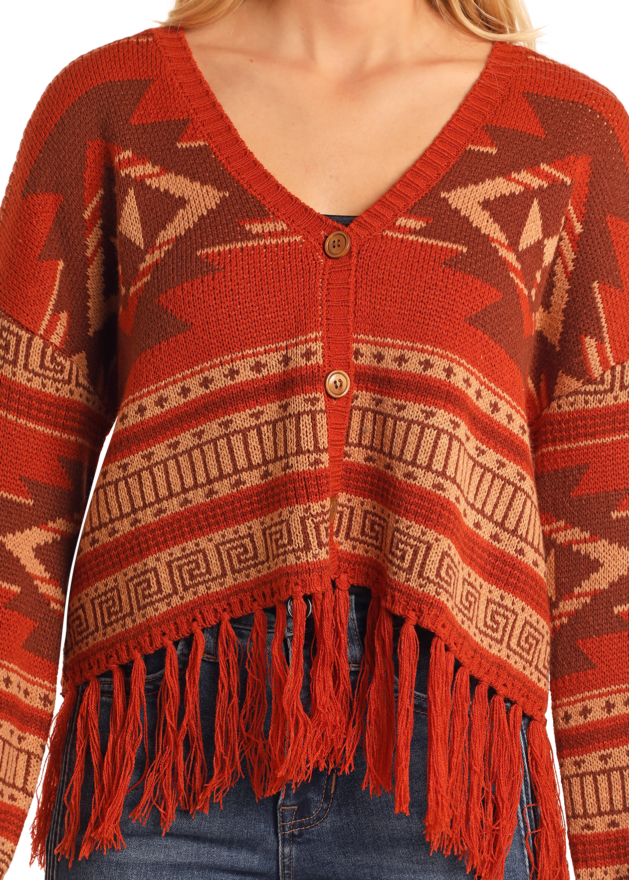 A woman wears the Rock & Roll Denim Aztec Fringe Steer Skull Cardigan Sweater, featuring rust-colored geometric patterns, wooden buttons, and long fringe along the hem, styled with blue jeans.
