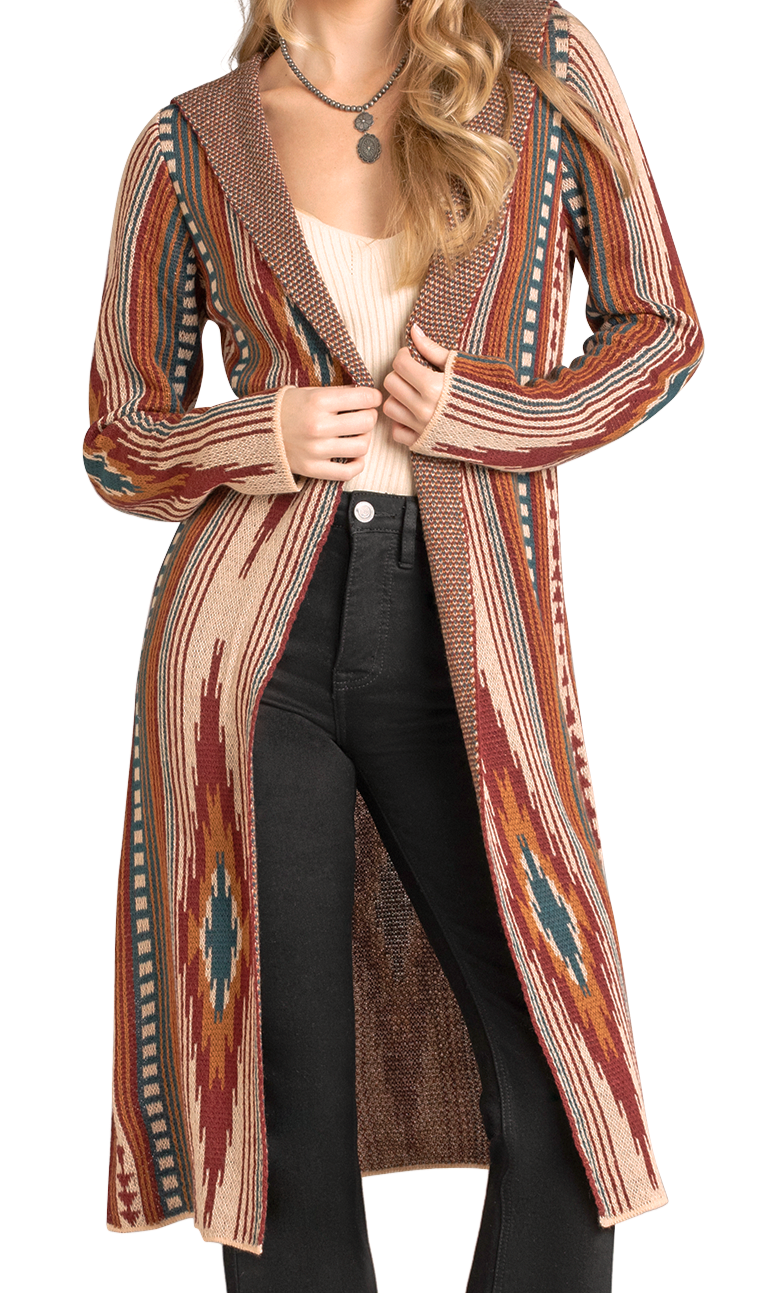 A woman styles the Rock & Roll Denim Aztec Hooded Knit Duster, featuring a vibrant southwestern geometric design, over a white top and black high-waisted jeans. She holds the open front near the collar and accessorizes with layered necklaces.