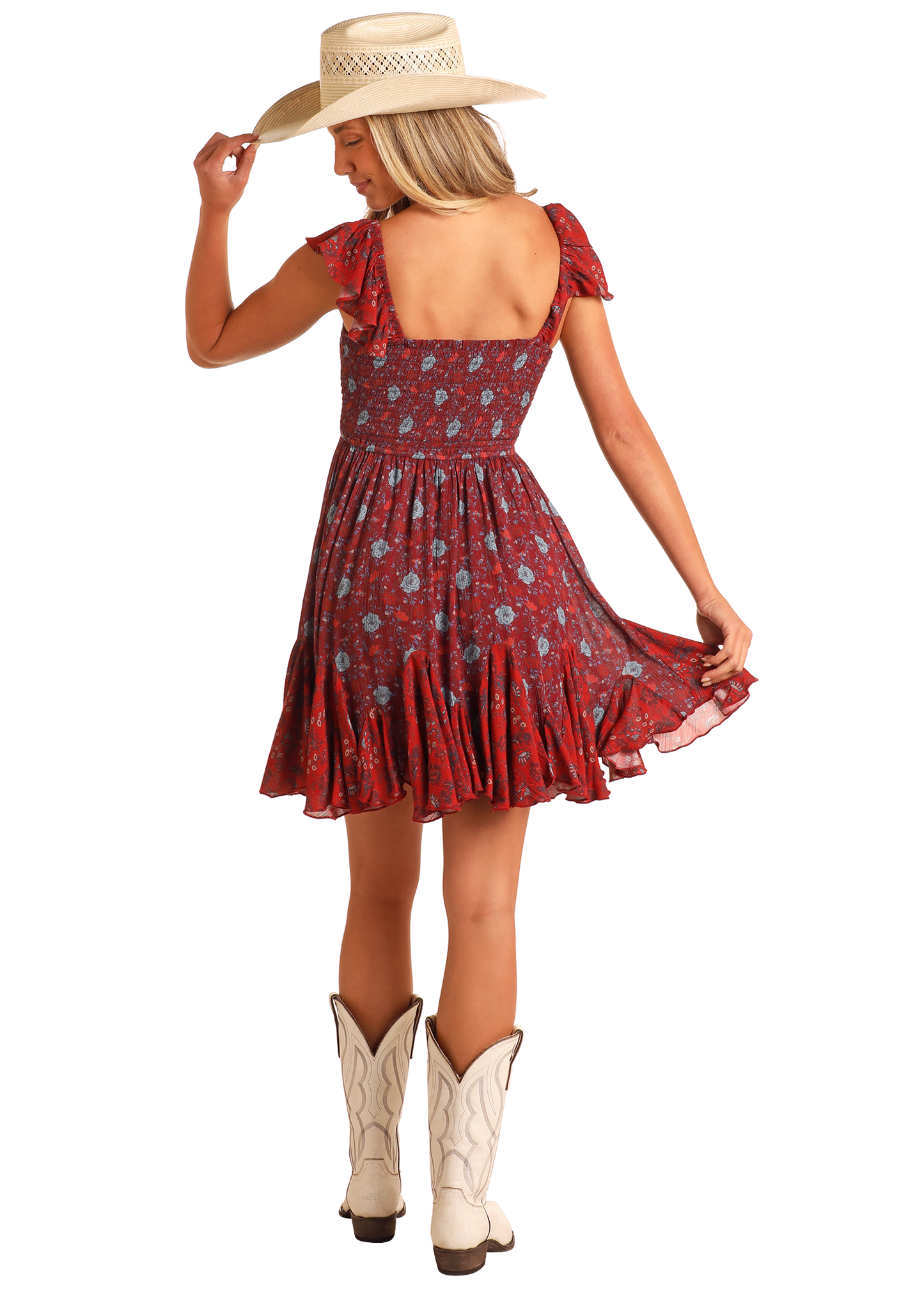 A woman in the Rock & Roll Denim Floral Godet Mini Dress, paired with white cowboy boots and a straw hat, stands with her back to the camera, holding her hat’s brim and slightly lifting her dress's skirt.