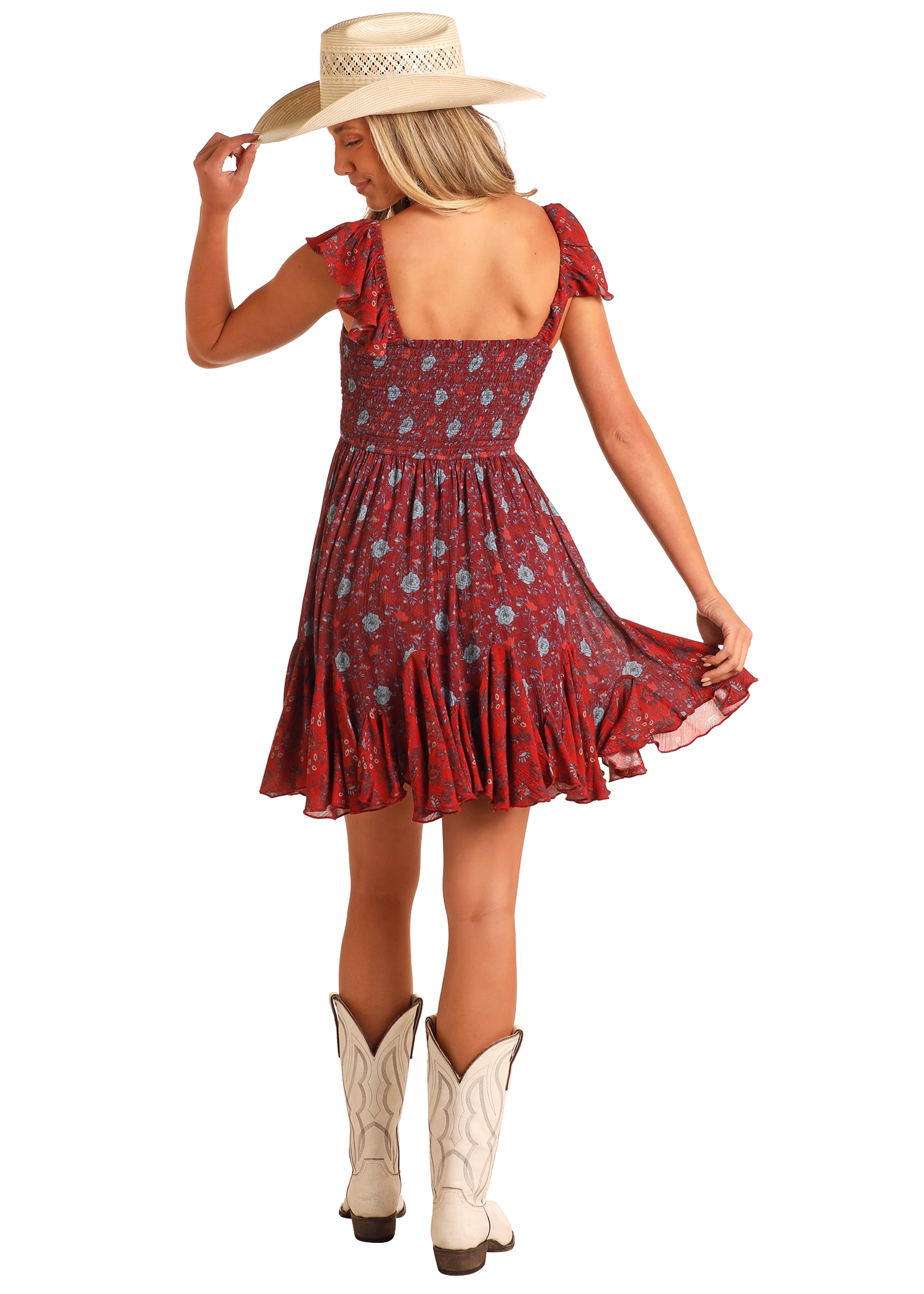 A woman in the Rock & Roll Denim Floral Godet Mini Dress, paired with white cowboy boots and a straw hat, stands with her back to the camera, holding her hat’s brim and slightly lifting her dress's skirt.