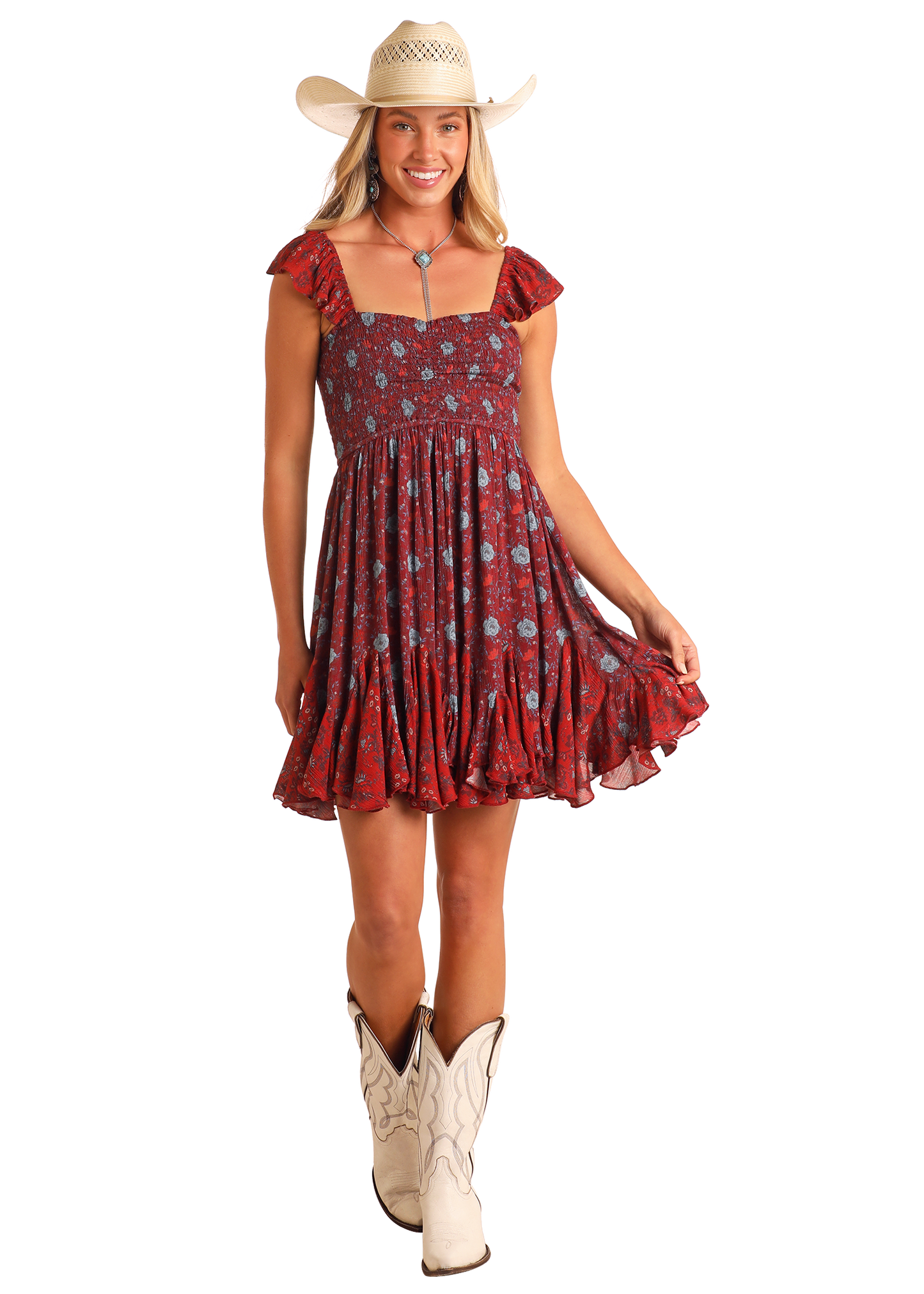 A smiling woman in a white cowboy hat, Floral Godet Mini Dress by Rock & Roll Denim with ruffled sleeves and hem, and white cowboy boots stands against a plain background, holding the edge of her dress.