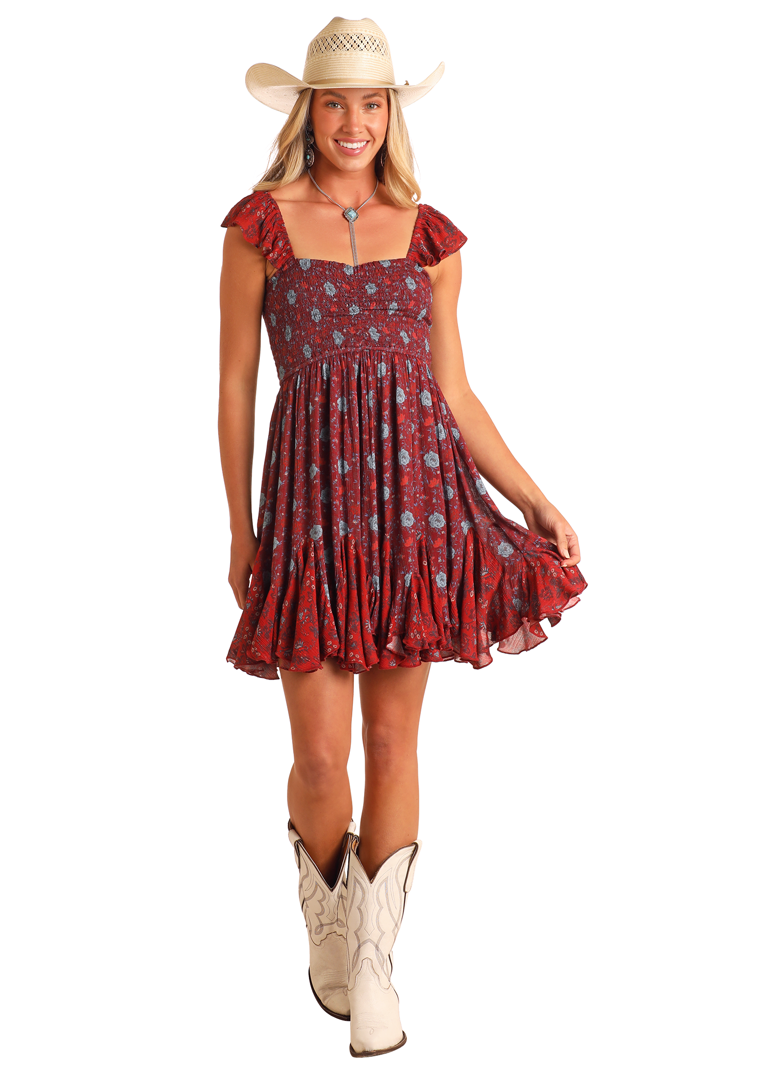 A smiling woman in a white cowboy hat, Floral Godet Mini Dress by Rock & Roll Denim with ruffled sleeves and hem, and white cowboy boots stands against a plain background, holding the edge of her dress.