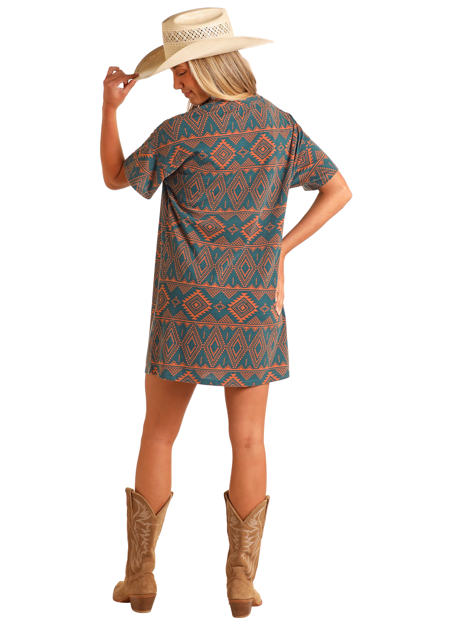 A woman in a Rock & Roll Denim Aztec Print T-Shirt Dress, wide-brimmed straw hat, and brown cowboy boots stands with her back to the camera, holding her hat’s brim with one hand.