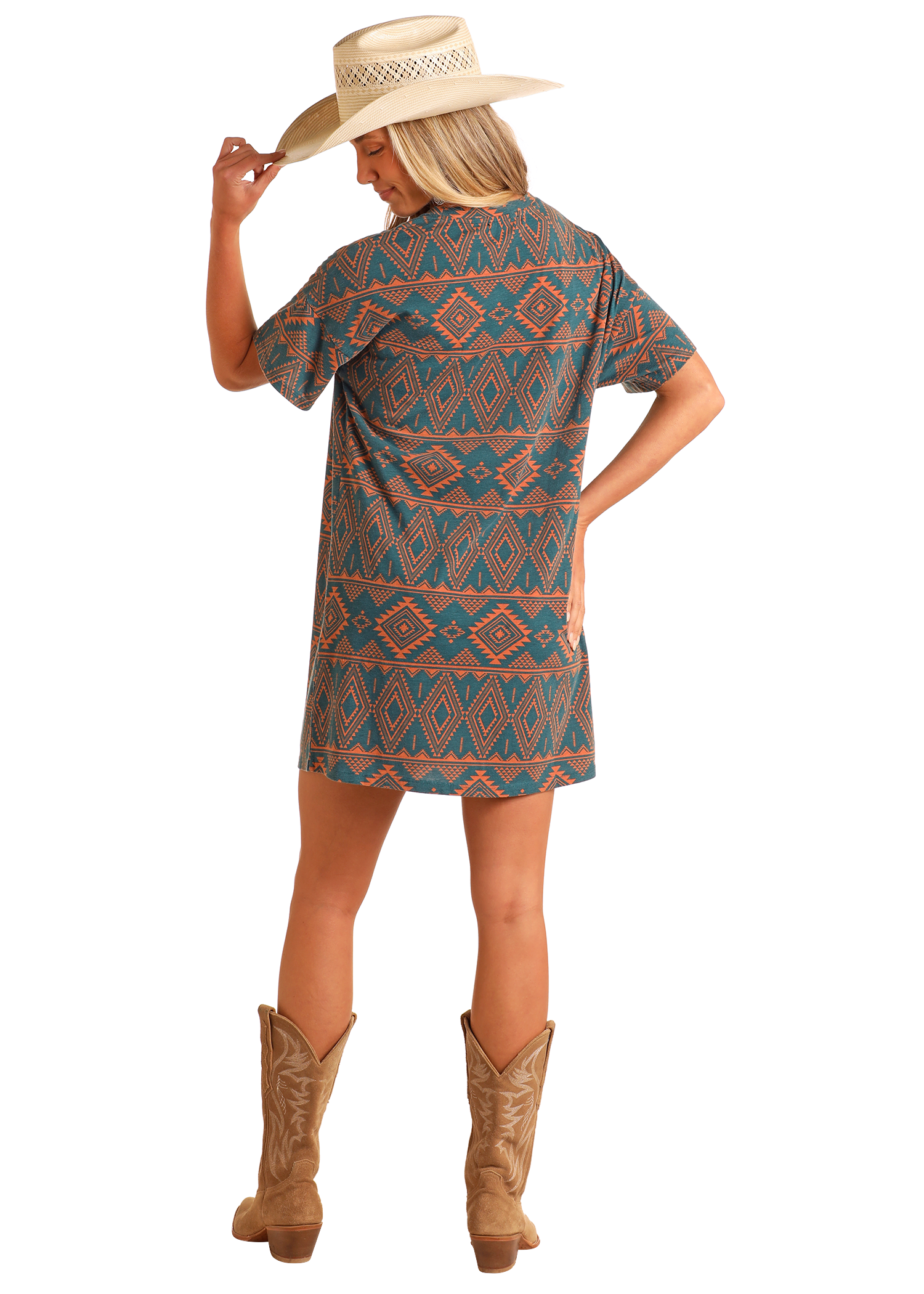 A woman in a Rock & Roll Denim Aztec Print T-Shirt Dress, wide-brimmed straw hat, and brown cowboy boots stands with her back to the camera, holding her hat’s brim with one hand.