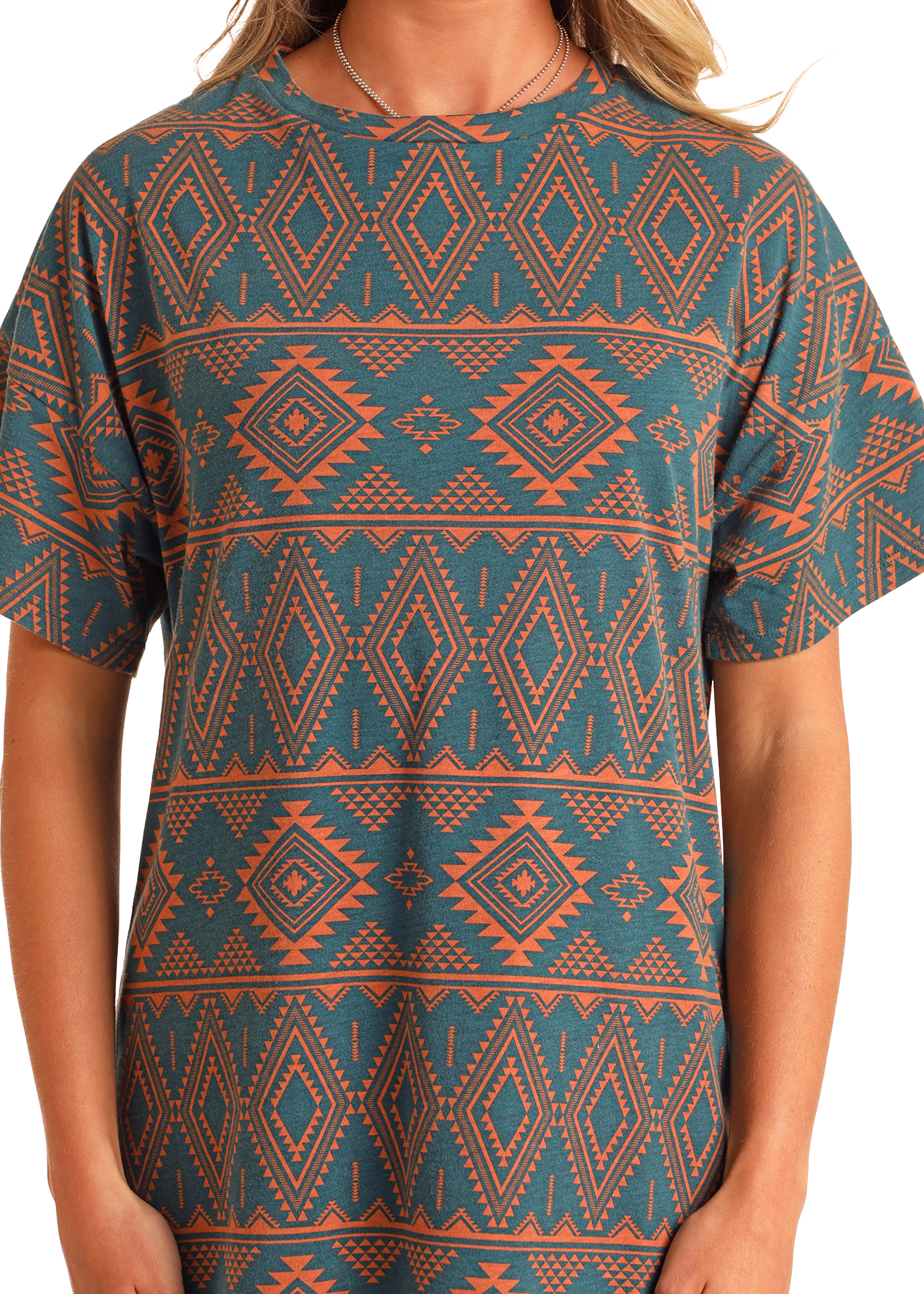 A person wears the Rock & Roll Denim Aztec Print T-Shirt Dress, a teal cotton dress with short sleeves and an orange geometric and diamond Aztec pattern. Only the torso and part of the arms are visible.